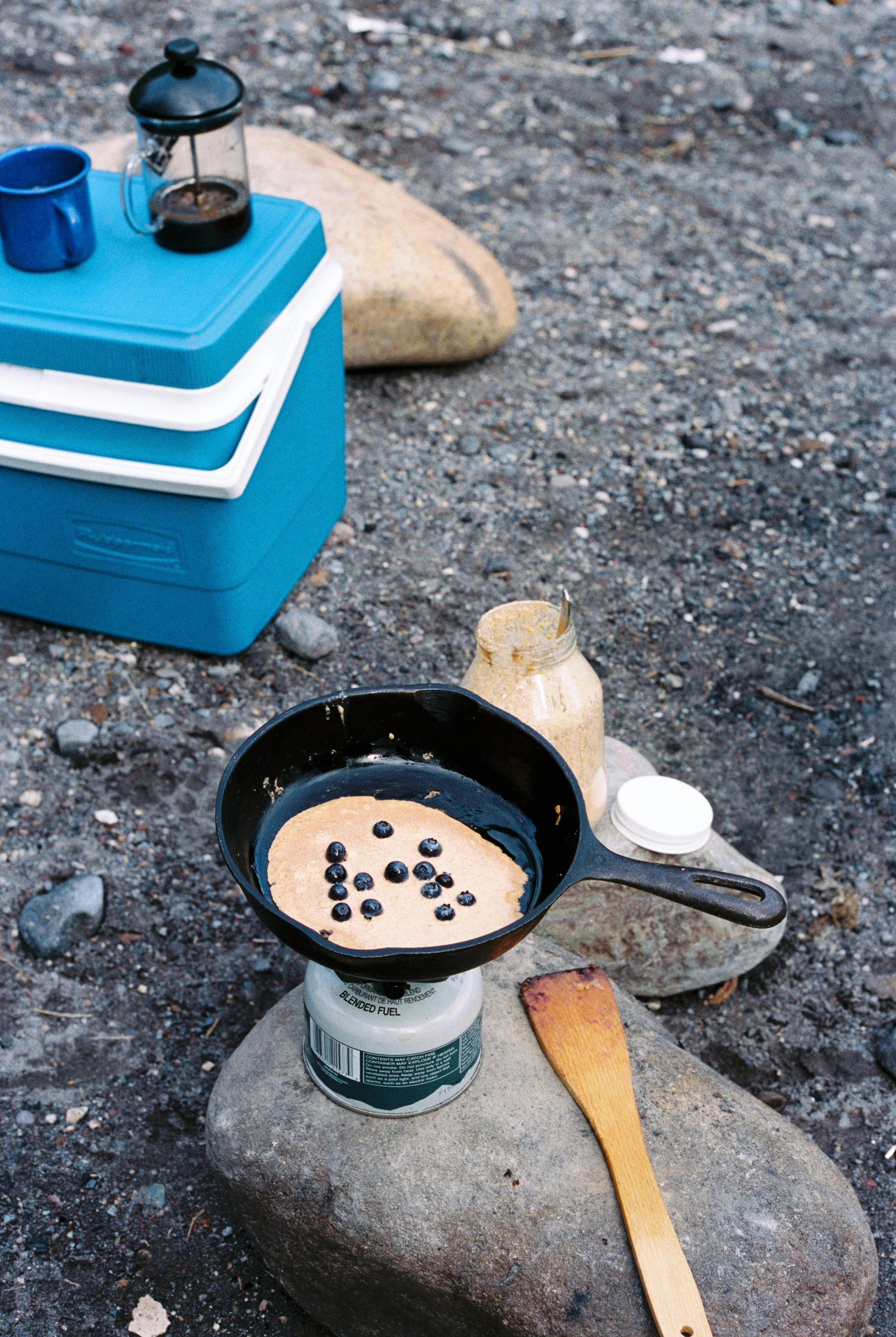 A camping stove on a rock cooking pancakes with blueberries, surrounded by a spoon, a jar of syrup, and other camping supplies on the ground outdoors.