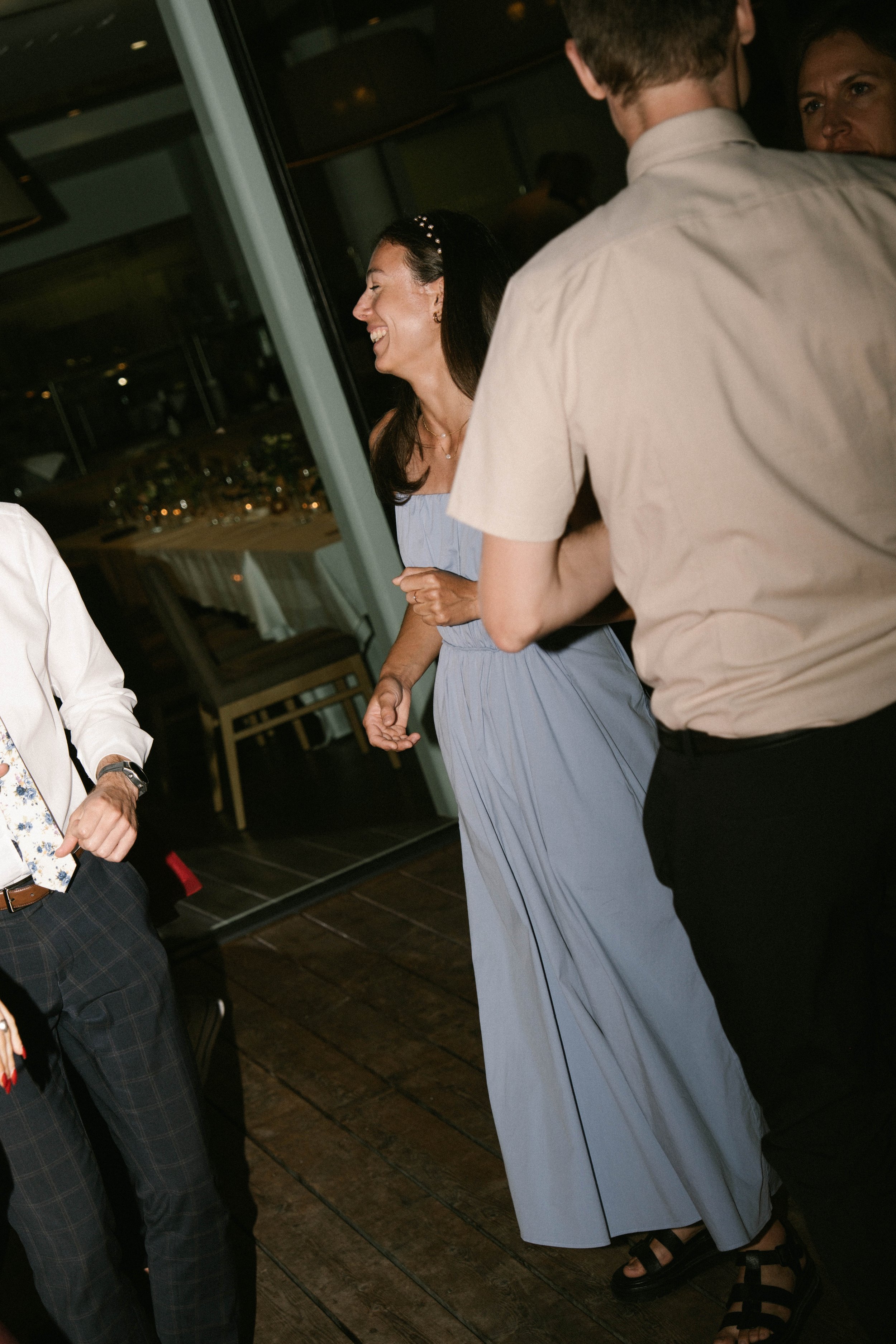 Group of people socializing at an indoor event, with focus on a woman laughing and wearing a light blue dress and a pearl headband, surrounded by others in semi-formal attire in Bend Oregon.