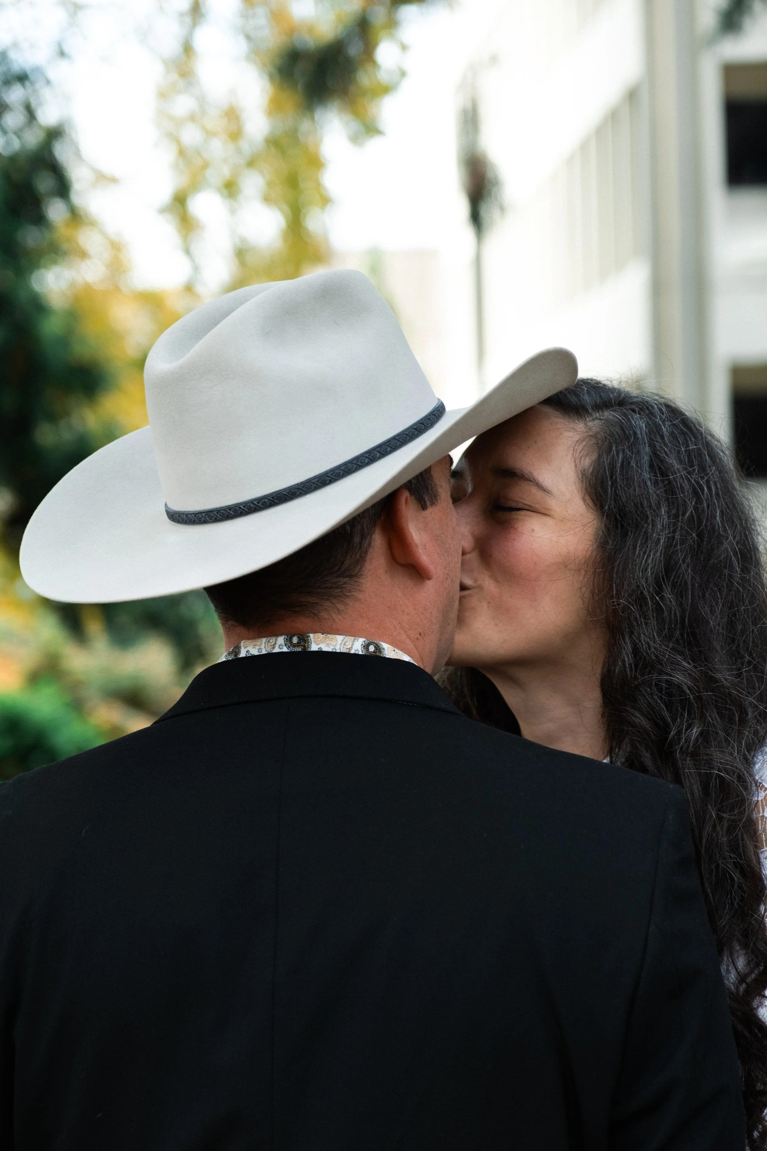 A man and a woman kissing outdoors, with the man wearing a white hat and a dark suit, and the woman with long dark curly hair and a white top.