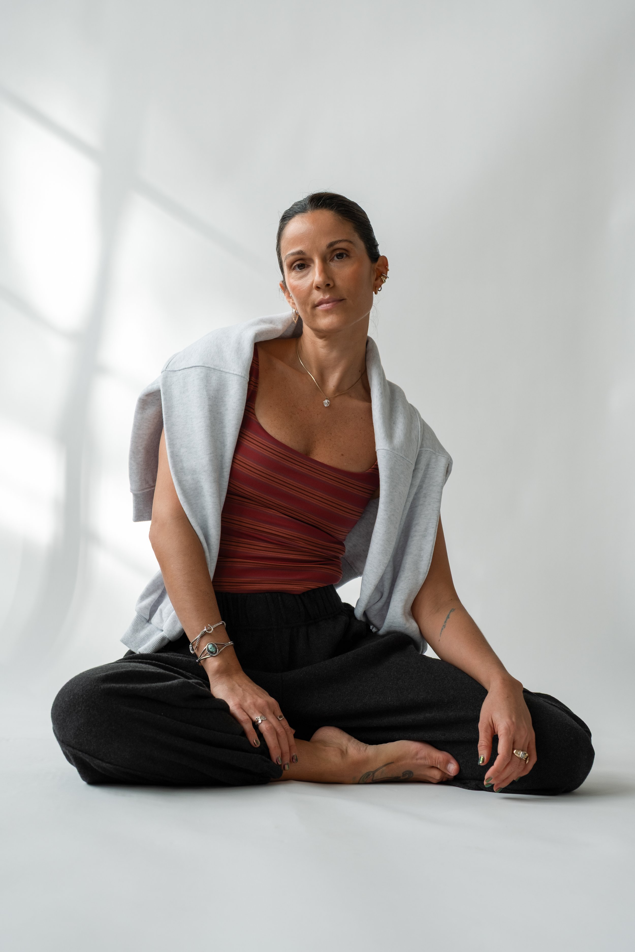 A woman sitting cross-legged on the floor in a bright, minimalist space. She is wearing a red striped top, black pants, and a gray hoodie draped over her shoulders, with multiple rings and bracelets.