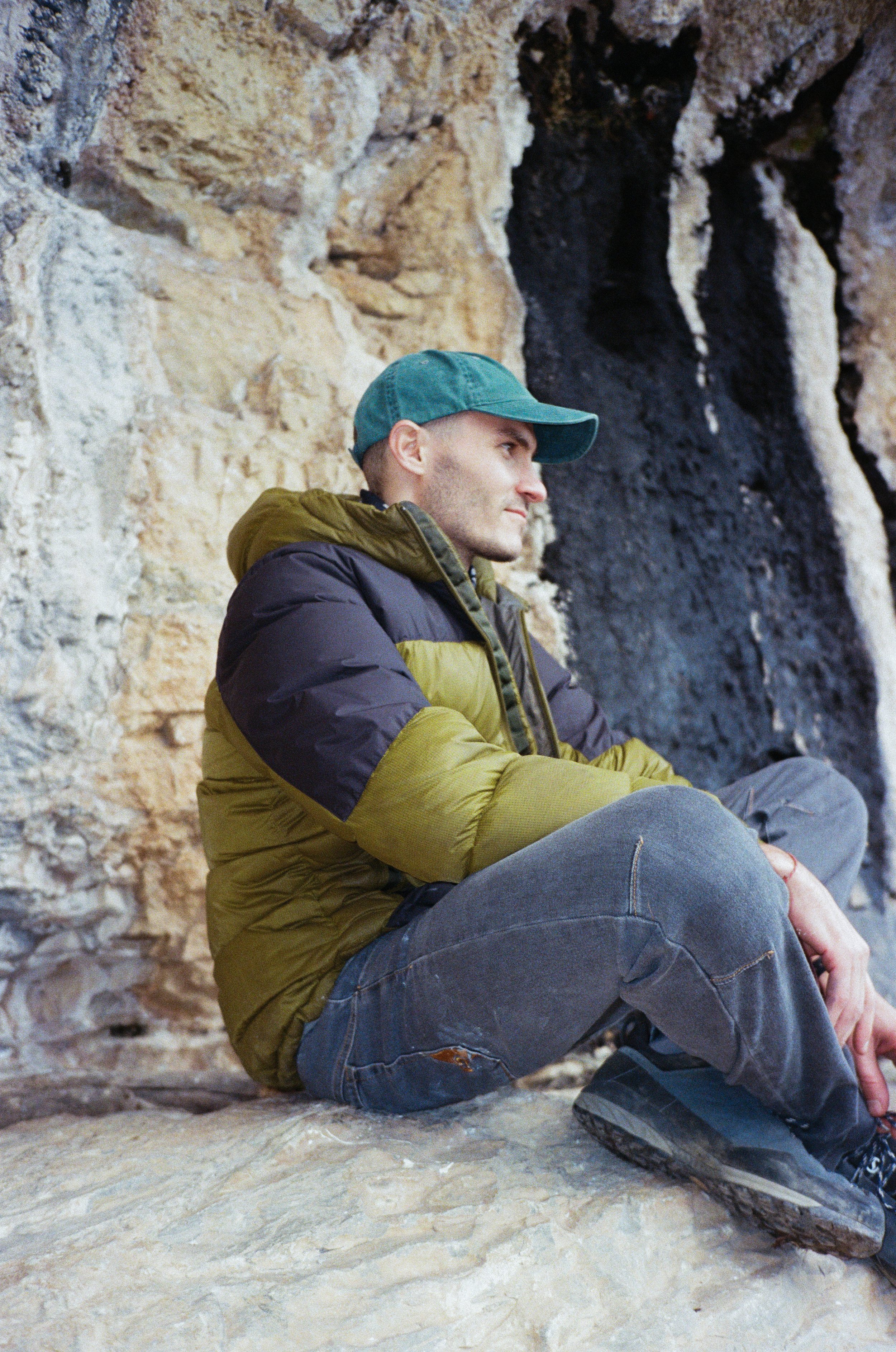 A man sitting on a rocky surface against a textured rock wall, wearing a green cap, brown and black puffy jacket, gray pants, and outdoor shoes, looking thoughtfully to the side.