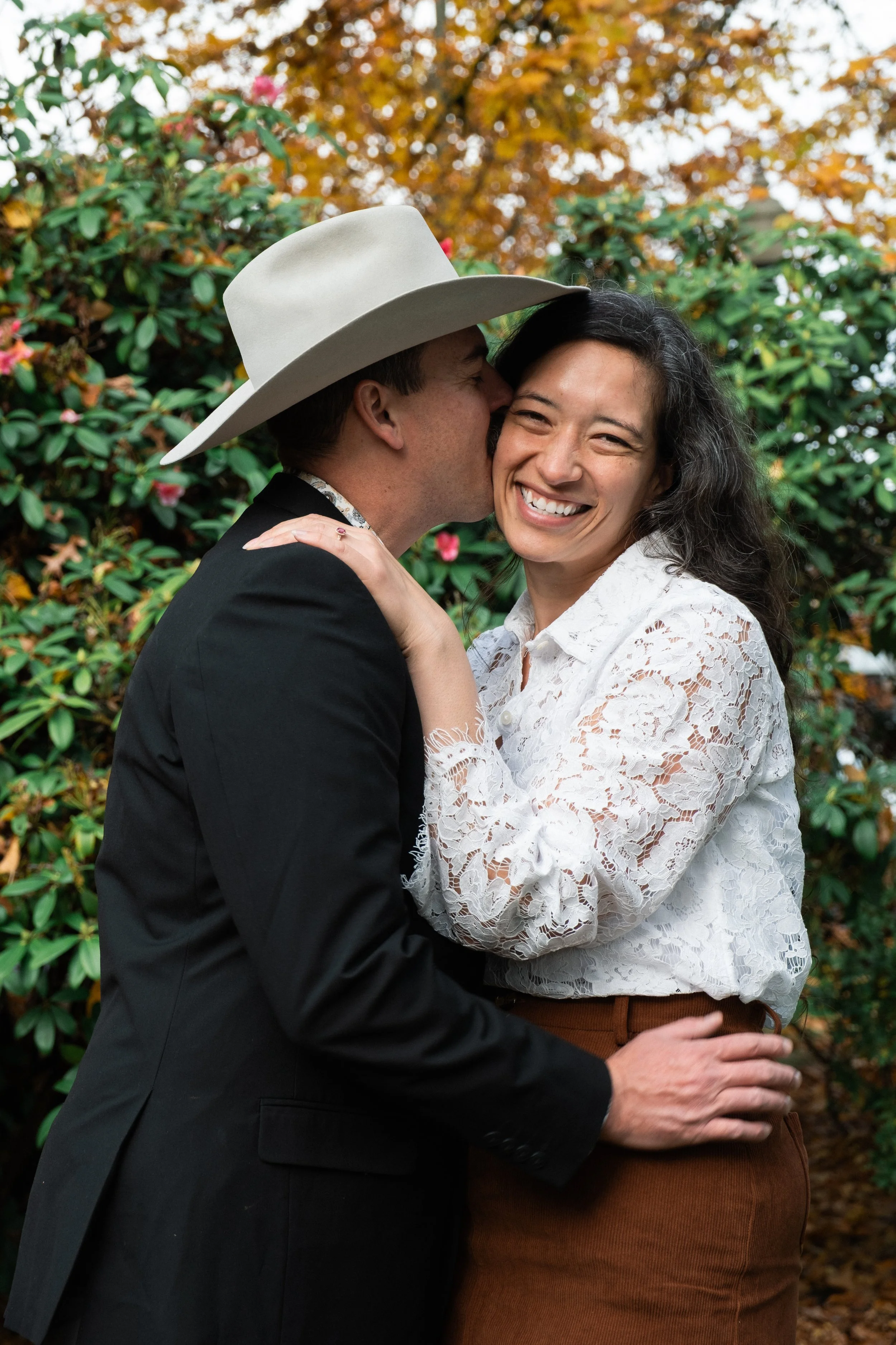 A man wearing a large white cowboy hat kisses a woman on the cheek while she smiles, standing in front of greenery with autumn leaves.
