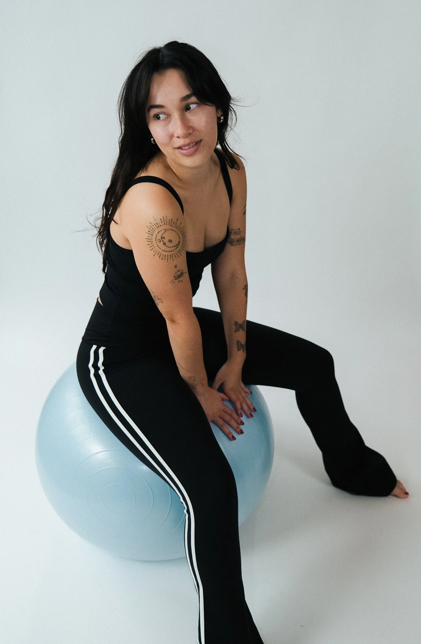 A woman with black hair and tattoos sitting on a light blue exercise ball in a white room, wearing a black tank top and black pants with white stripes.