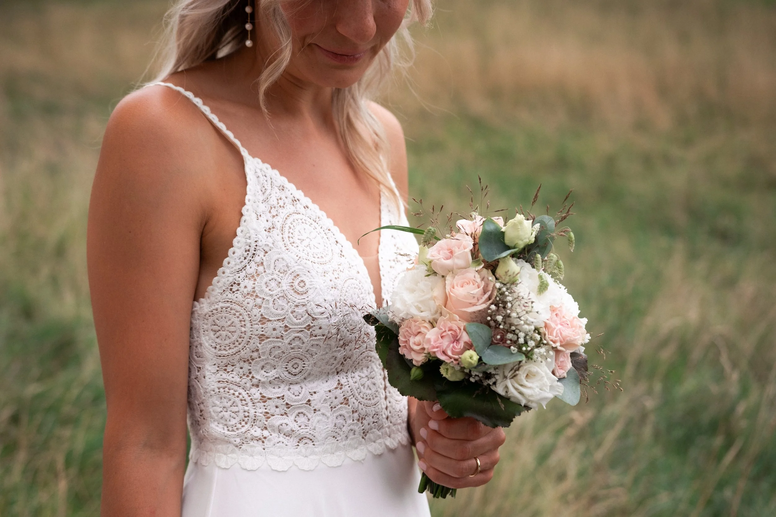 A woman in a white lace dress holding a bouquet of pink and white flowers outdoors in a grassy field.