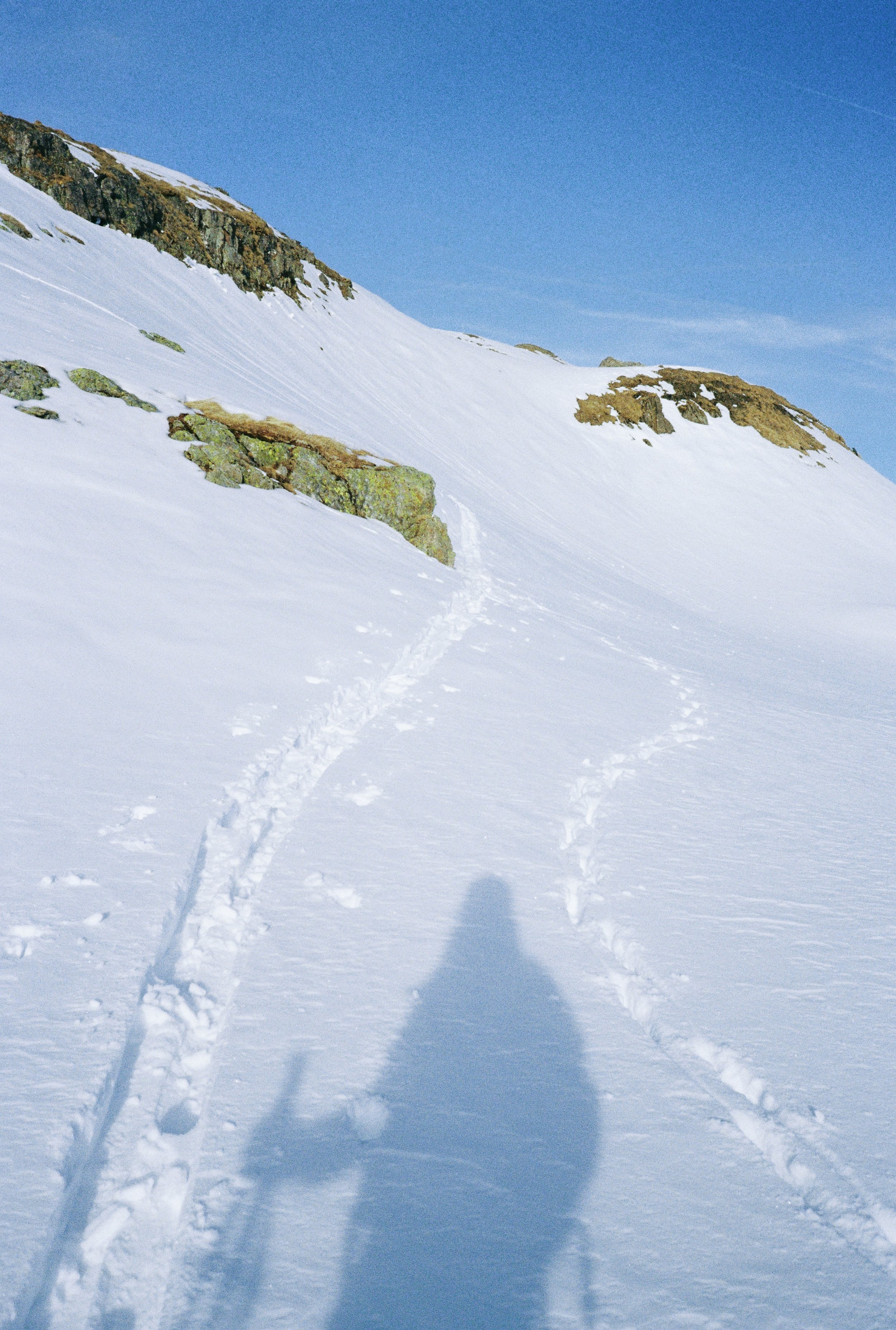 Snow-covered mountain slope with ski trail and rocky patches, shadow of a person taking the photo, and a clear blue sky.