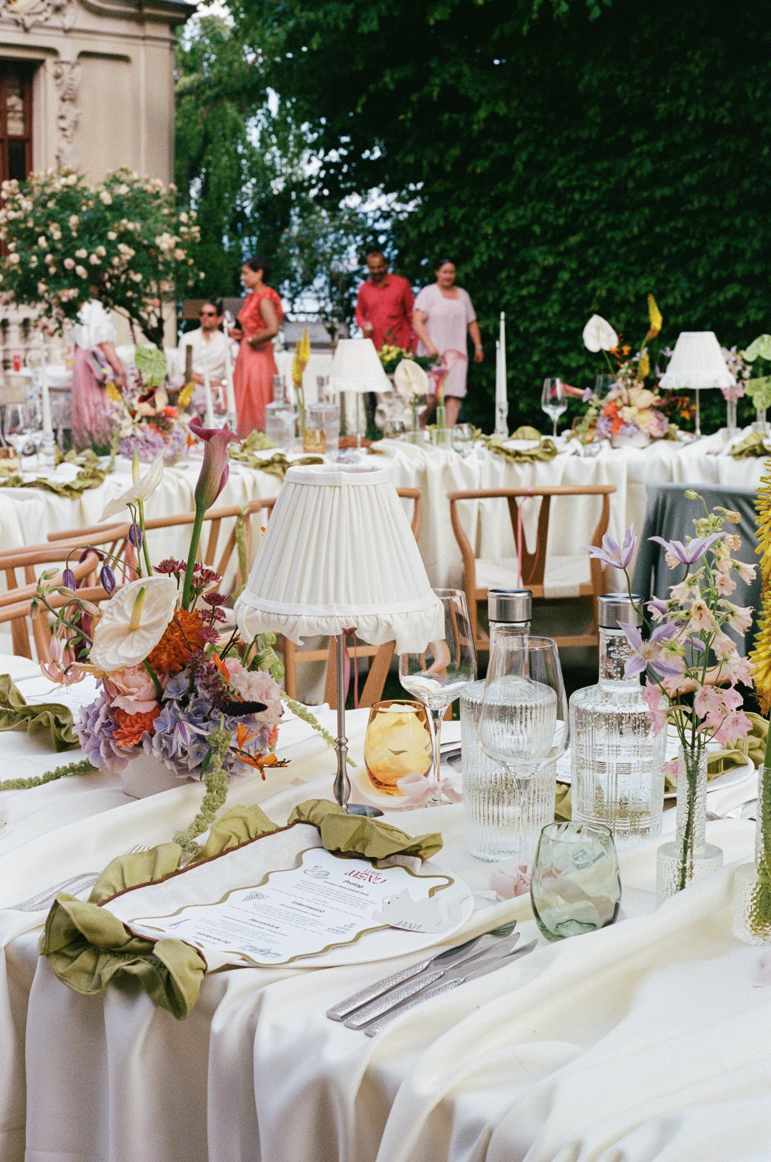 Outdoor banquet table set with flowers, wine glasses, water glasses, candles, and a menu, surrounded by greenery and trees, with a few people standing in the background near a building.