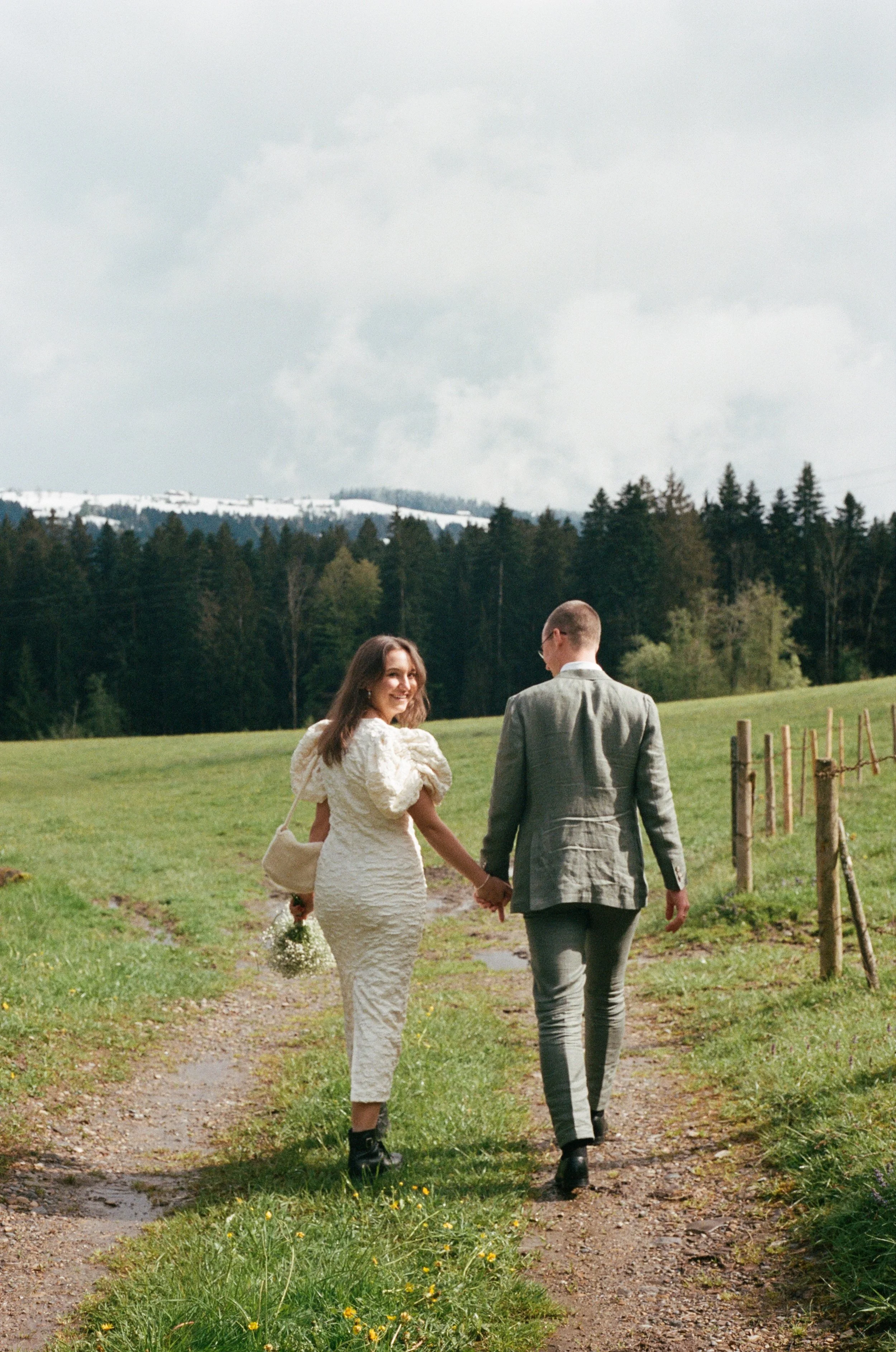 A smiling woman and a man holding hands and walking through a grassy field, with trees and a cloudy sky in the background.