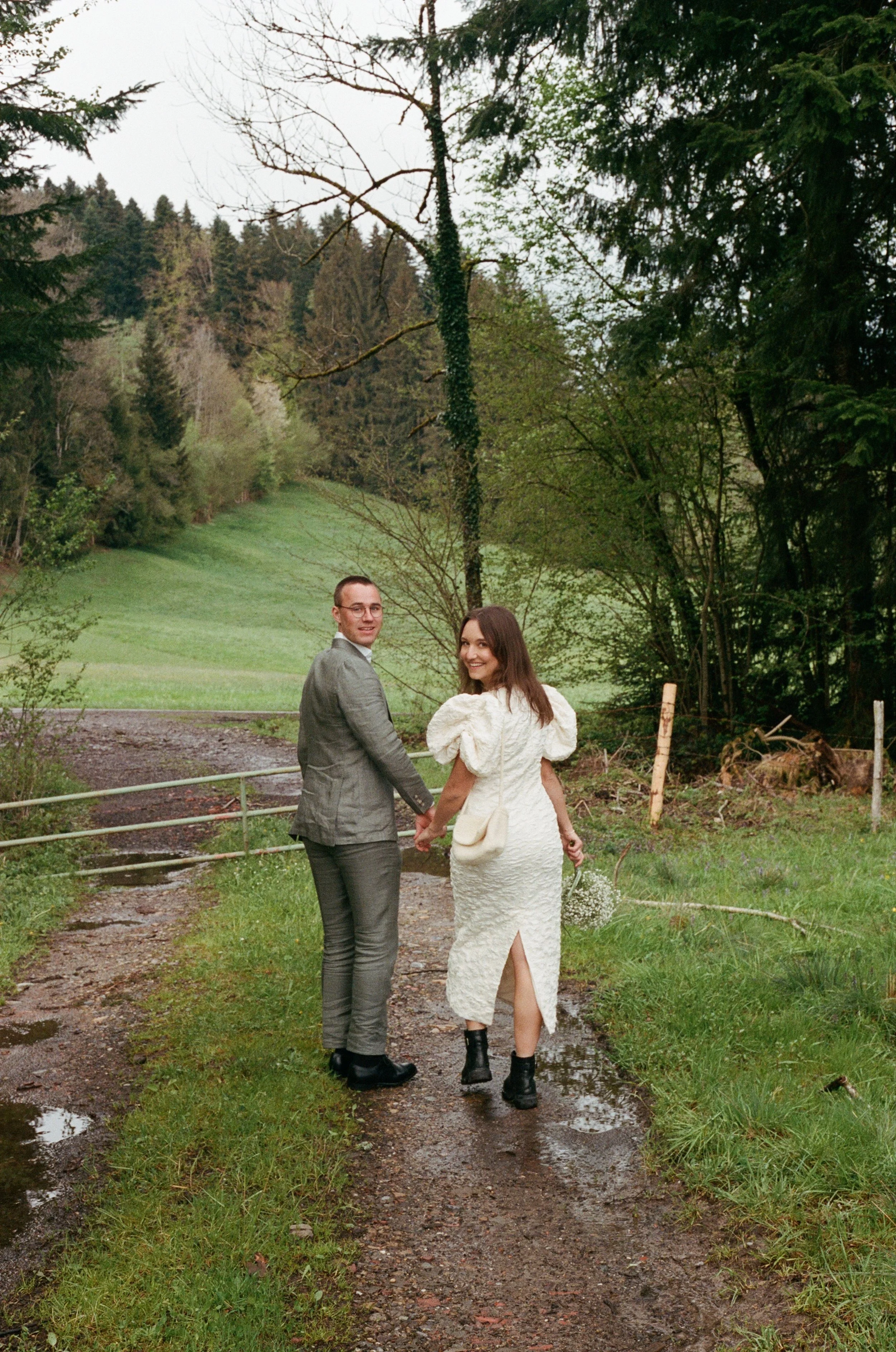 A couple holding hands and walking away on a muddy path in a lush green, wooded area, looking back at the camera with smiles.