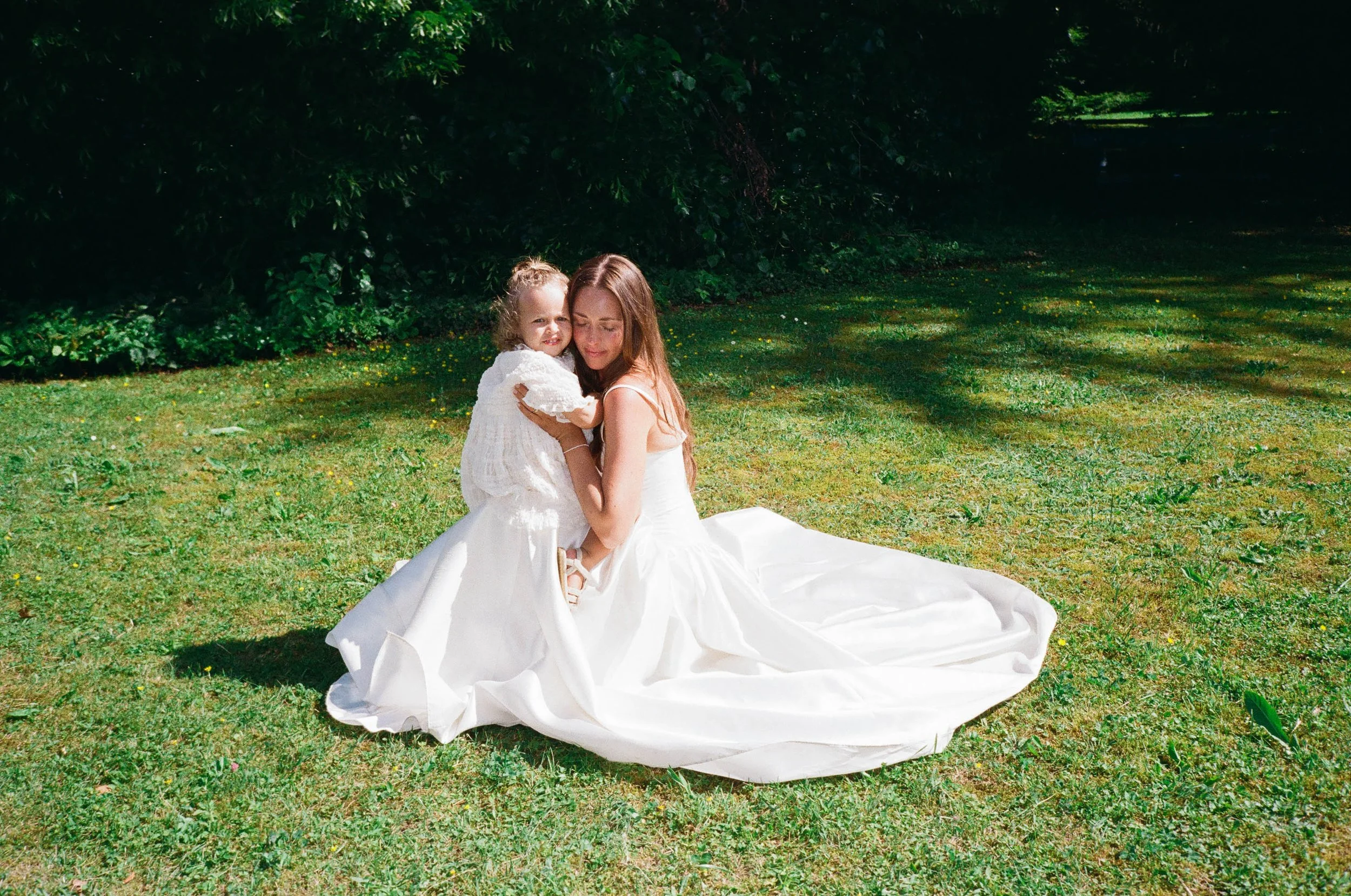 A bride and her daughter captured in a emotional and intimate moment at a scenic outdoor wedding on a grassy area with trees in the background, possibly for a wedding or special event in Switzerland. 