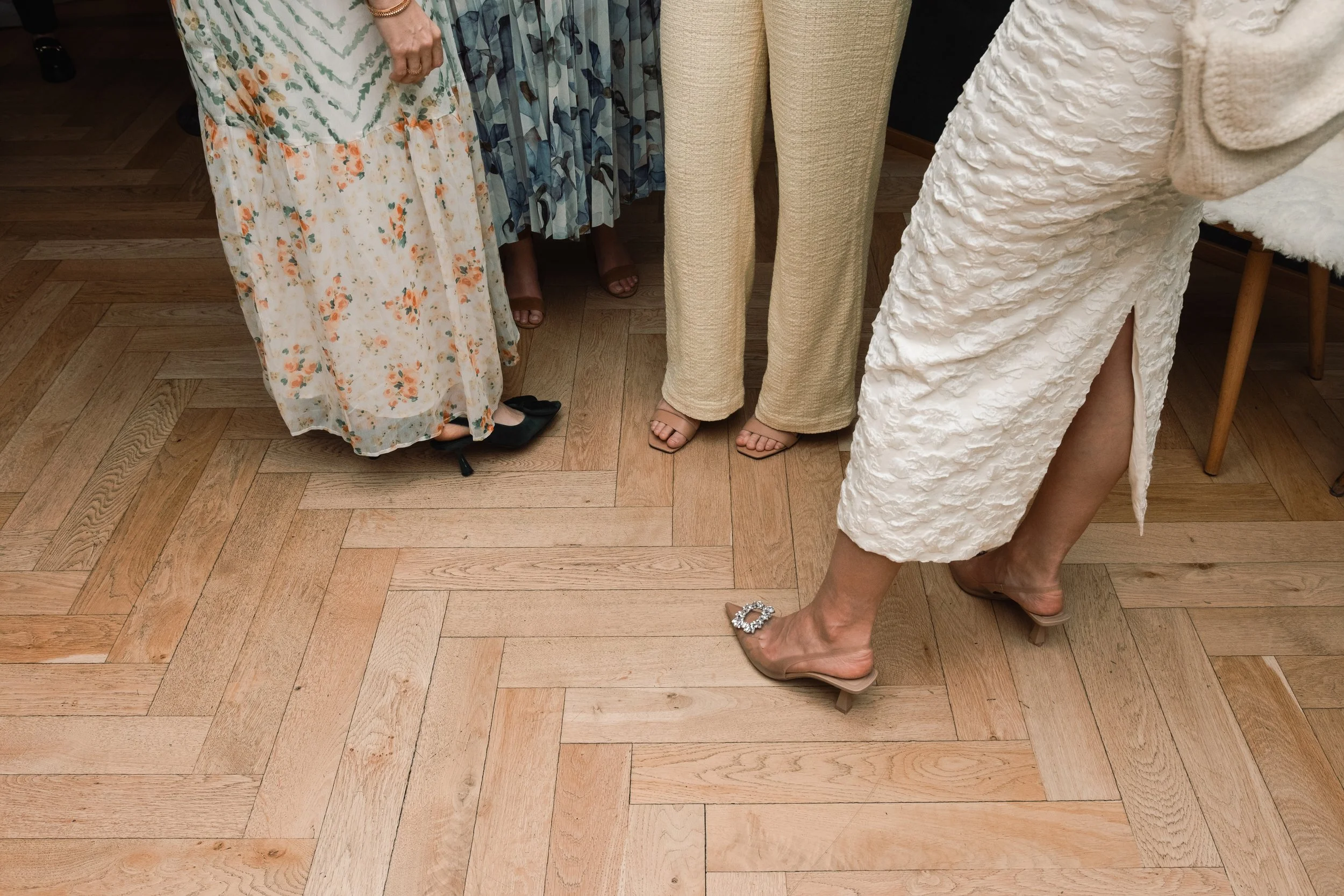 Part of a group of women standing on a wooden floor, wearing dresses and high heels.