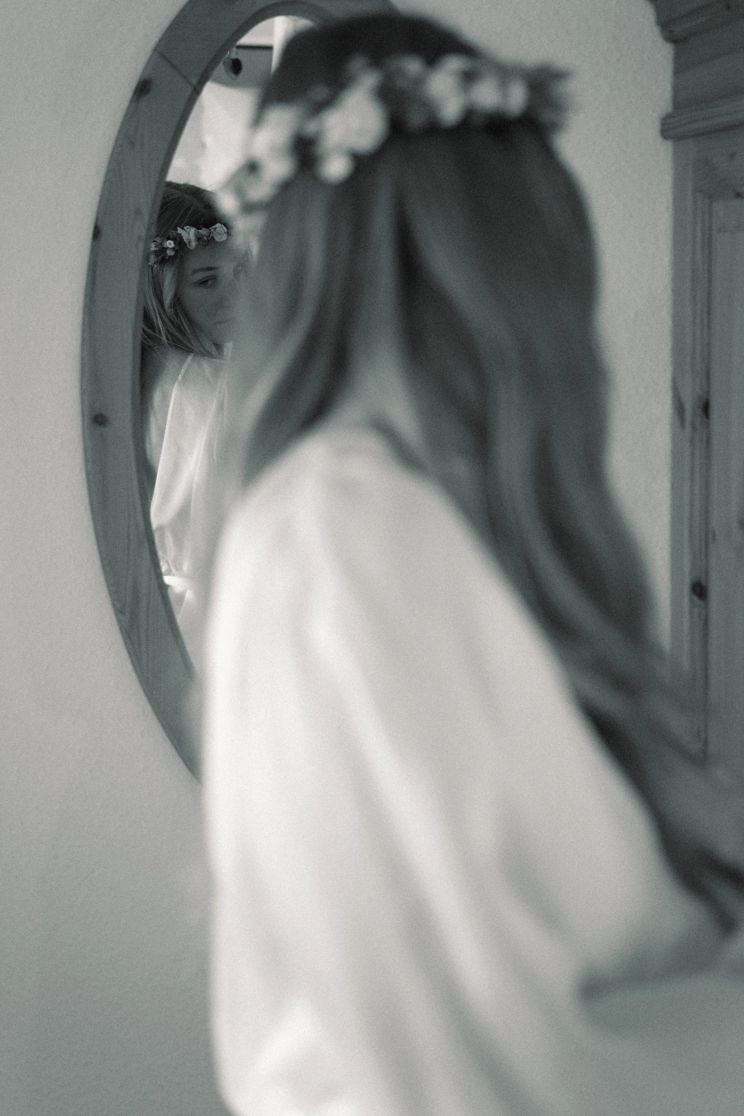 A woman with long hair and a floral headband looks at her reflection in an oval mirror, capturing her face in the reflection while her back faces the camera. The photo is in black and white.
