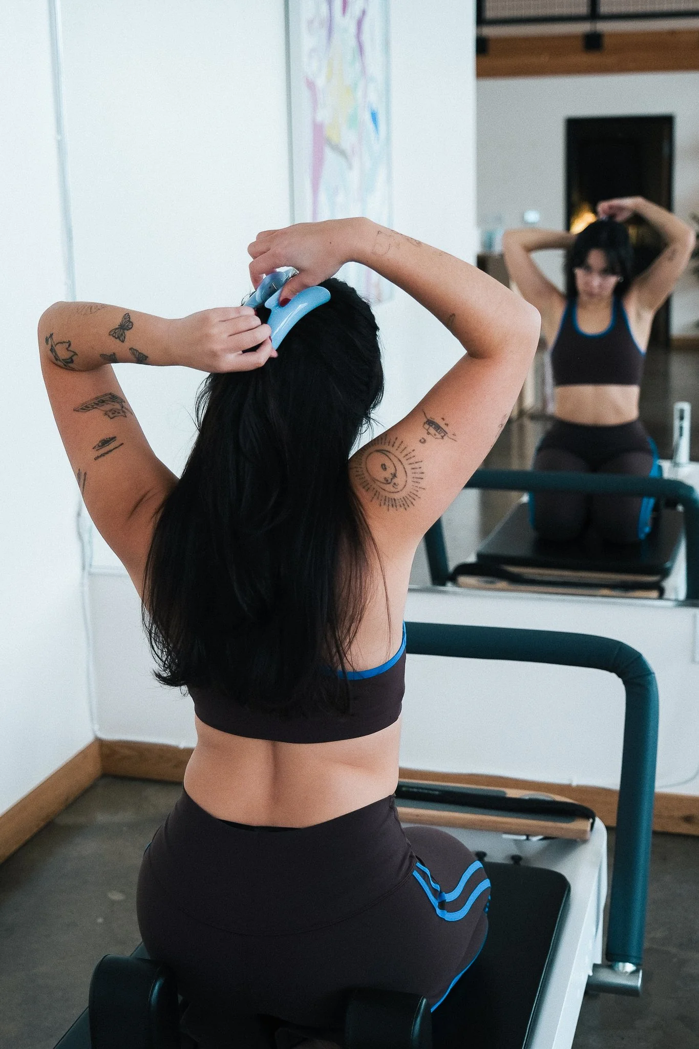 Two women exercising on a Pilates reformer machine in a fitness studio; one woman is stretching her arms behind her head, while the other is adjusting her hair in front of a mirror.
