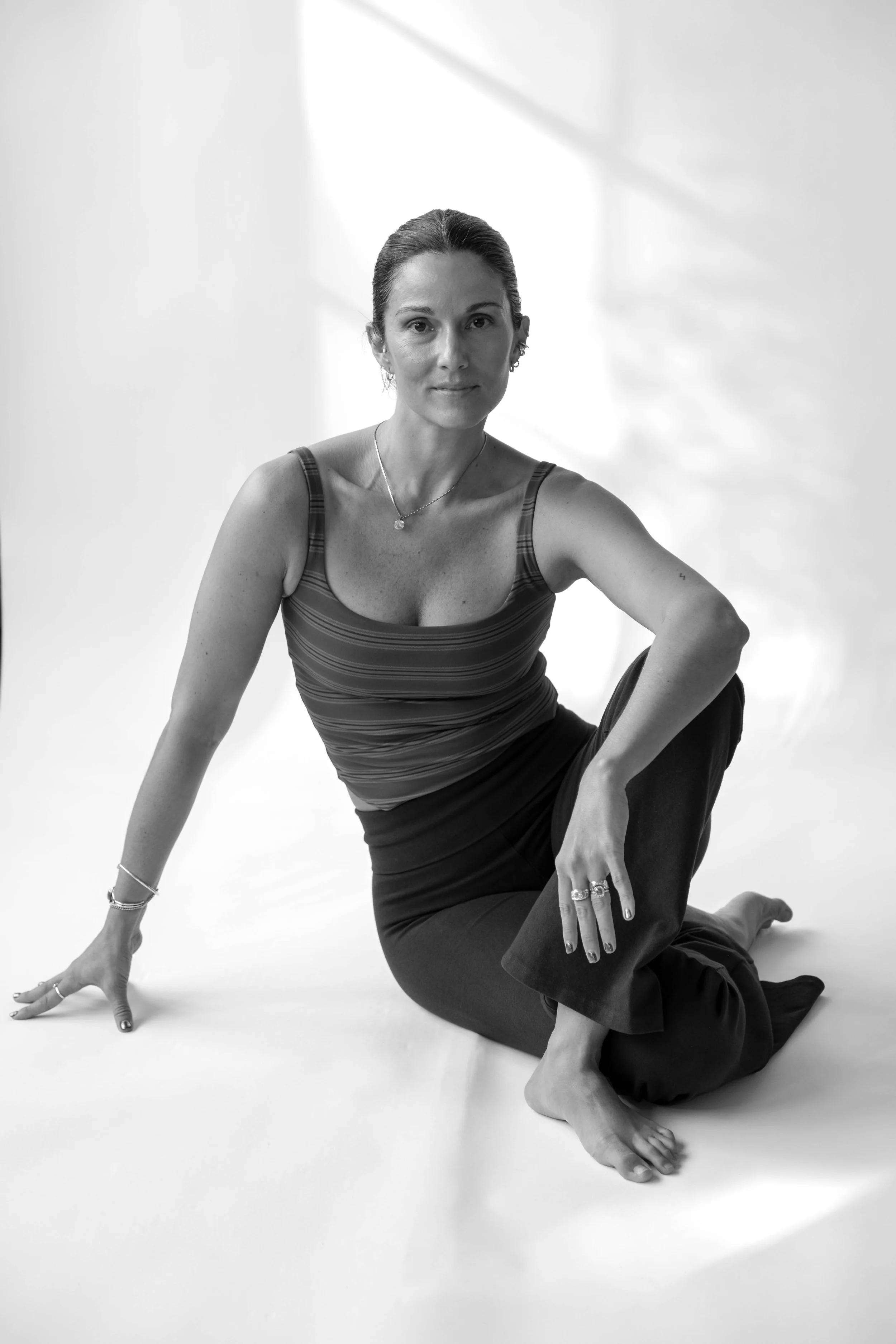 A woman sitting on the floor with one arm supporting her and one leg bent, wearing a striped sleeveless top and dark pants, posing in a studio with natural light.