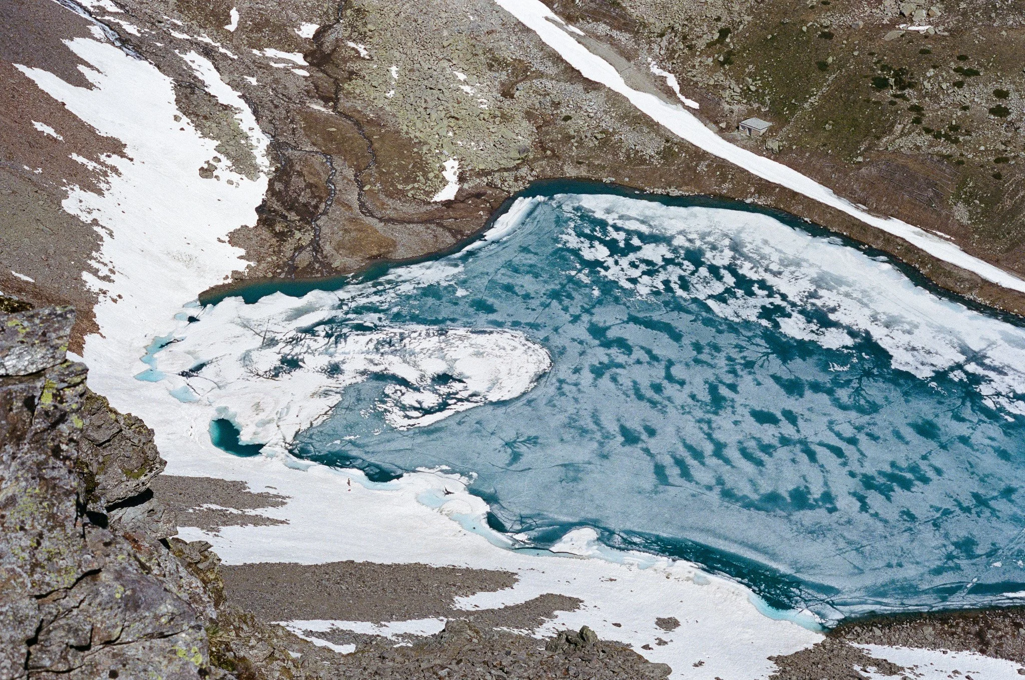 A partially frozen mountain lake surrounded by snow and rocky terrain, viewed from above.