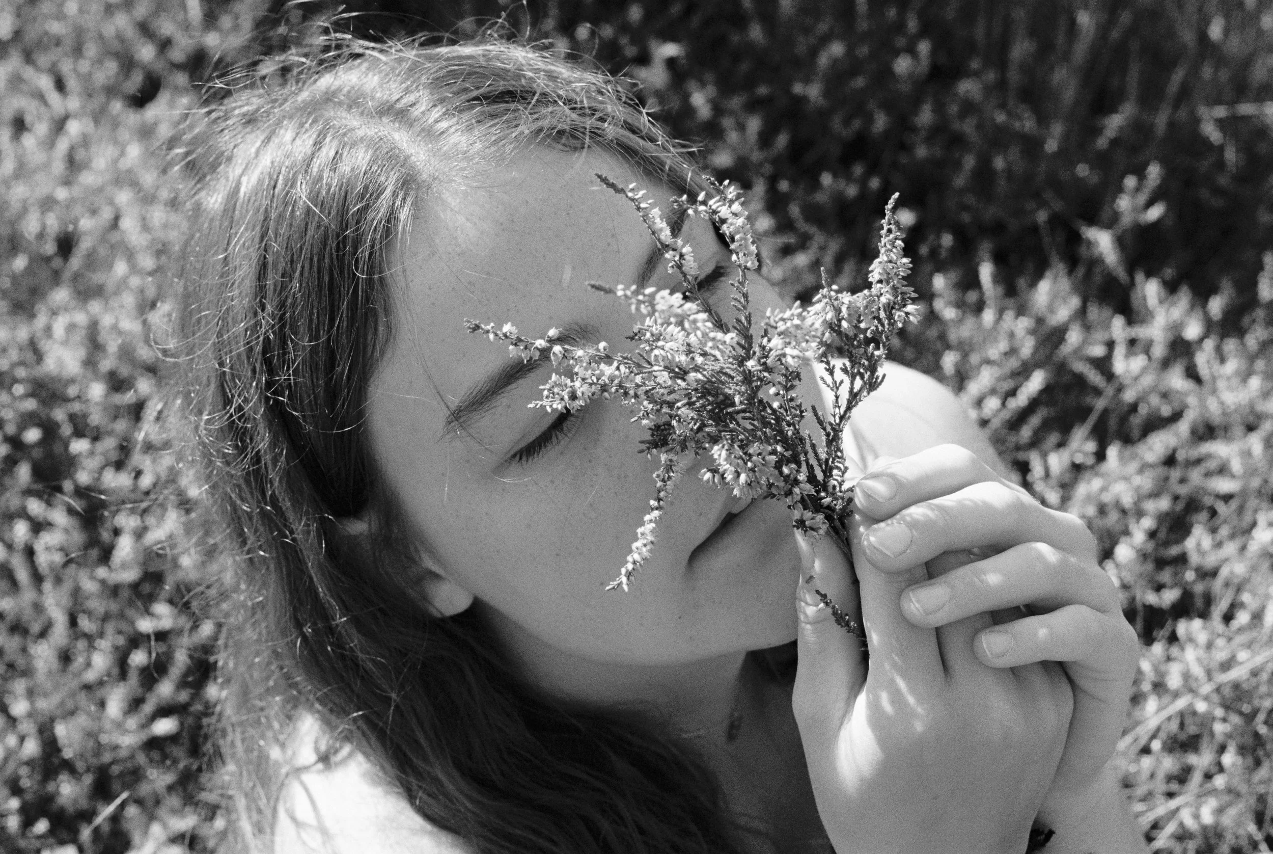 A black and white photo of a woman with closed eyes holding a plant close to her face outdoors.