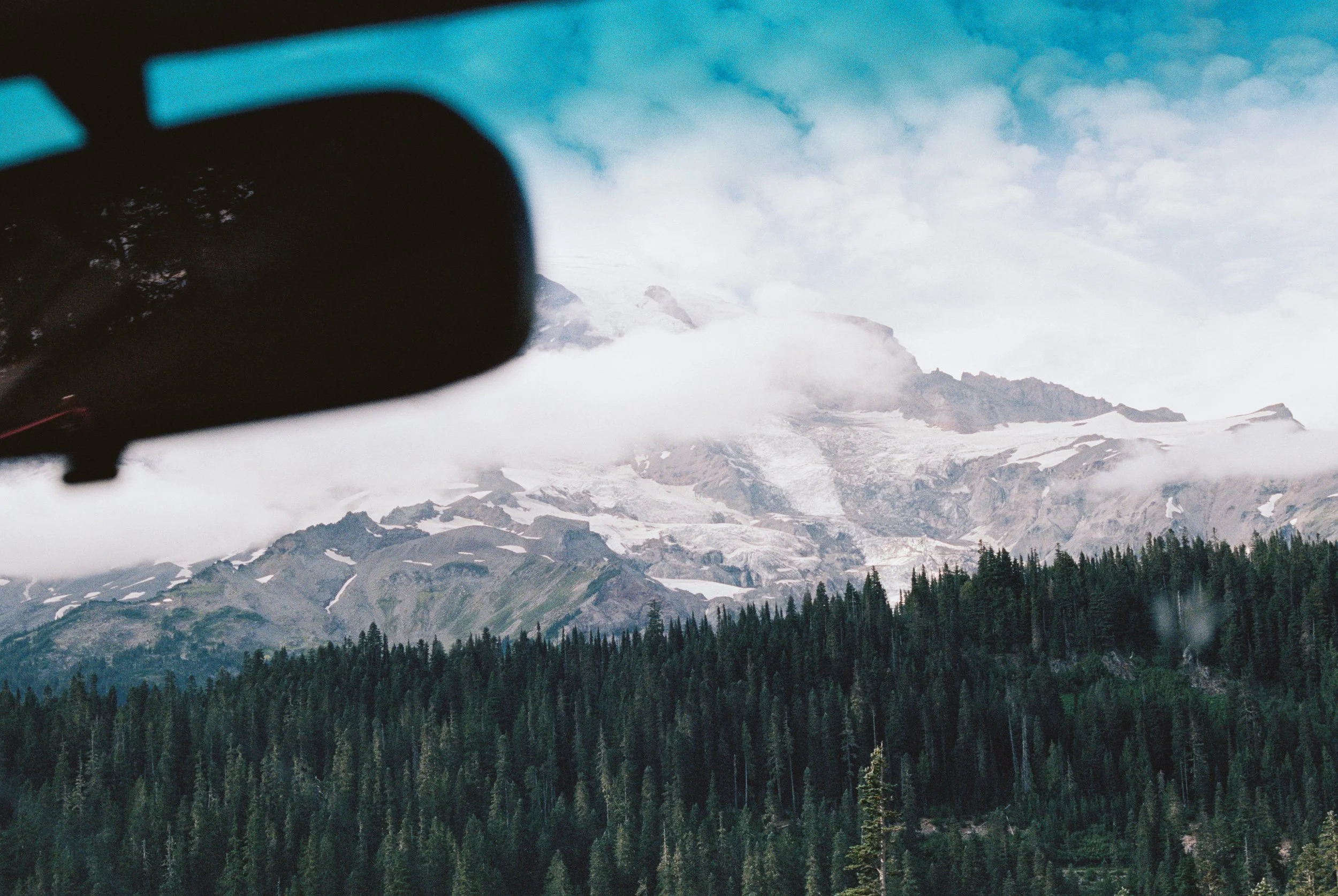 Mountain landscape with snow-capped peaks, dense dark green forest at the base, viewed partially through a car's side mirror and windshield.