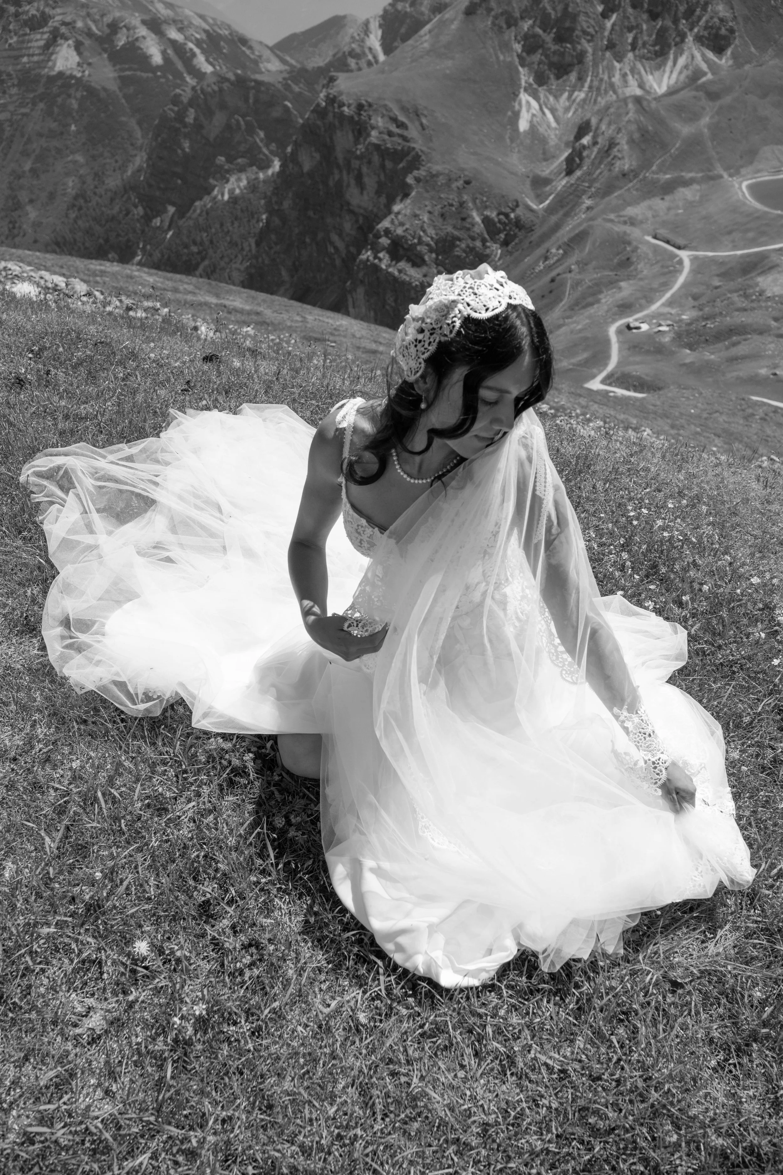A woman in a wedding dress sitting on grass in a scenic mountainous landscape, with a scenic background of mountains and a winding trail in Washington. 