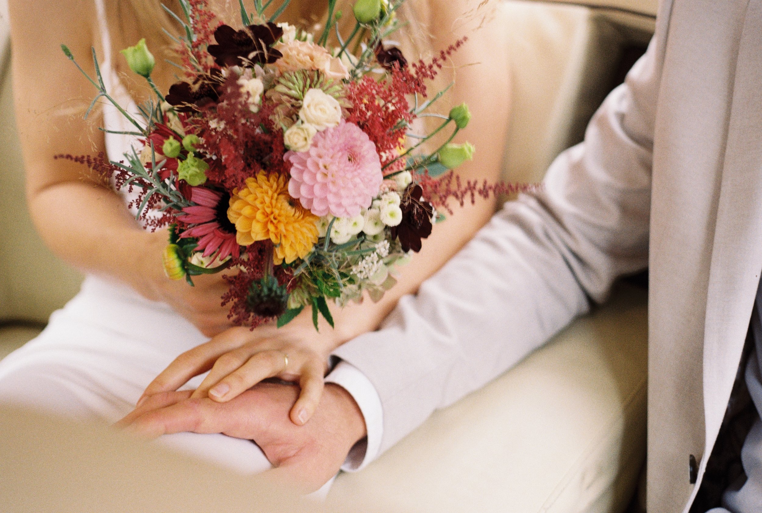 A person in a white suit holds a bouquet of colorful flowers, with their hand resting on their knee. The bouquet includes various pink, yellow, white, and dark-colored flowers.