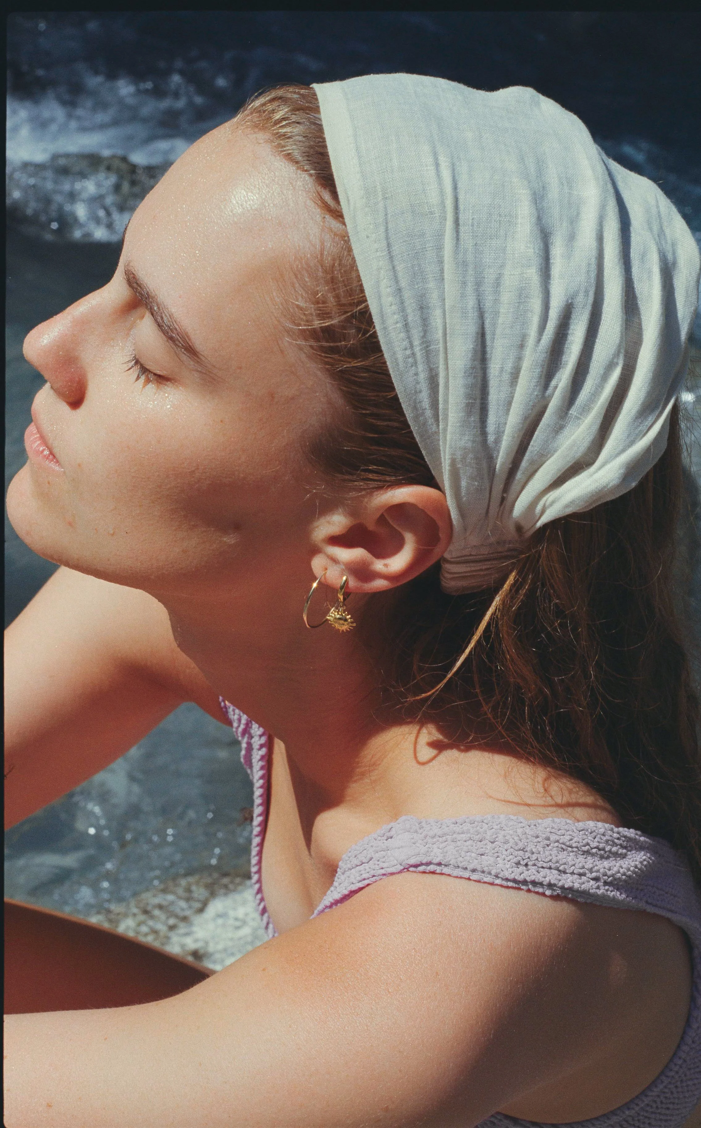 Close-up of a woman with her eyes closed, wearing a white headscarf and gold earrings, enjoying a sunny day near water.