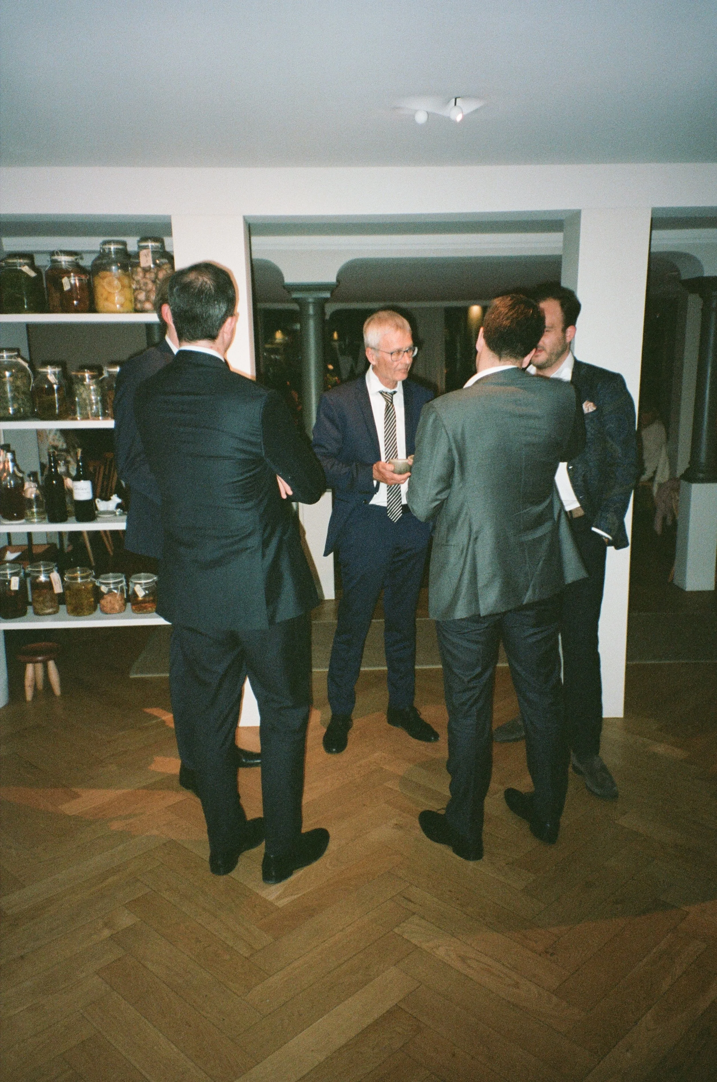 Five men in suits engaged in conversation at an indoor gathering or party, with shelves of jars and bottles in the background.