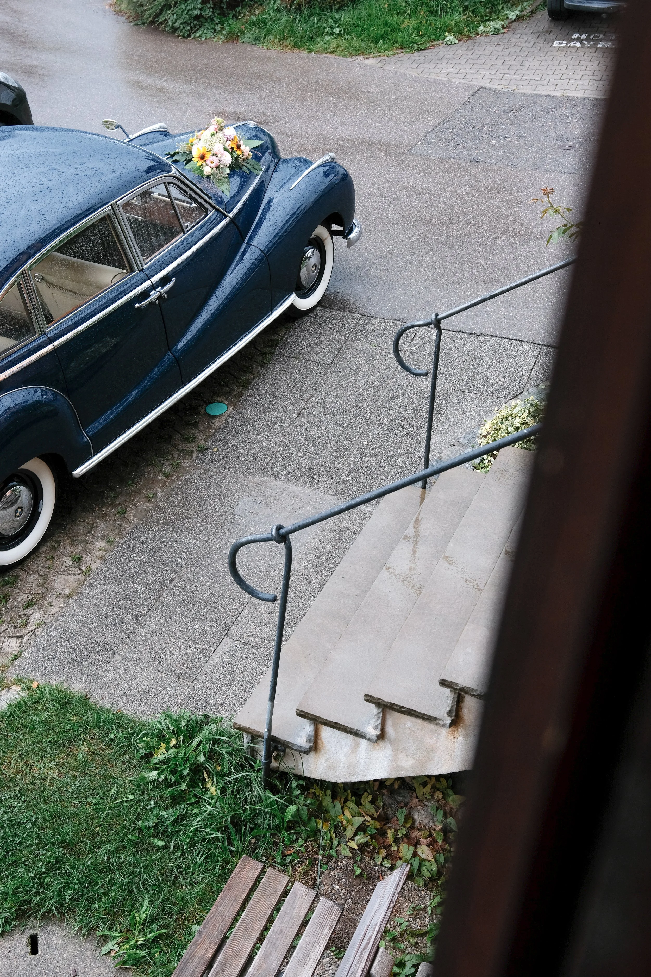 A vintage blue car with rain droplets on it, decorated with a bouquet of flowers on the hood, parked outside near a staircase leading to a house.