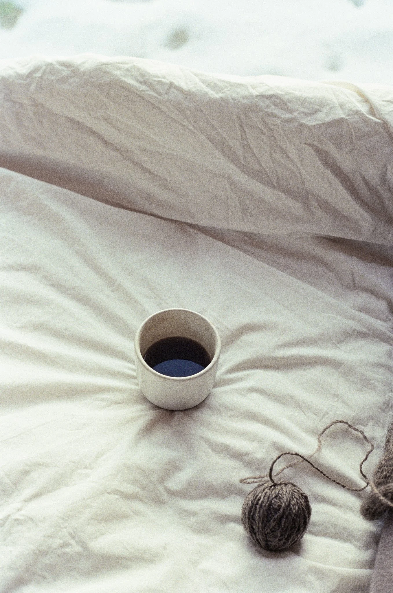 A white cup filled with black coffee resting on white bed sheets, with a ball of yarn and a knitting needle nearby.