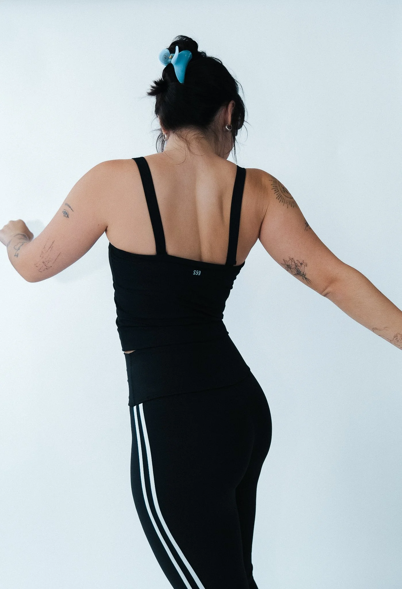 A woman with tattoos on her arms seen from the back, wearing a black tank top and black leggings with white stripes, and a blue hair clip.