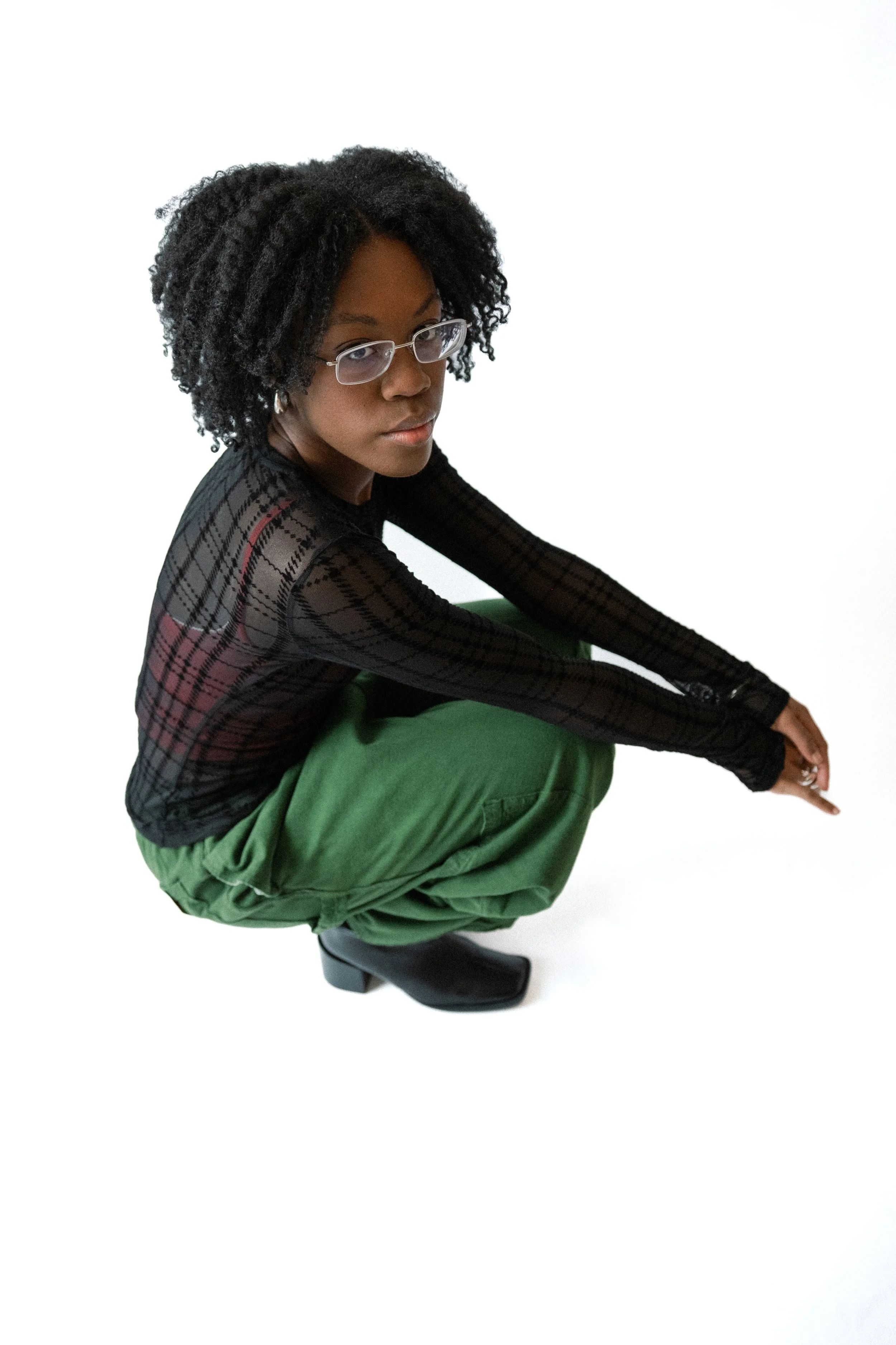 A woman squatting down with glasses, wearing a black sheer top and green pants, against a white background.