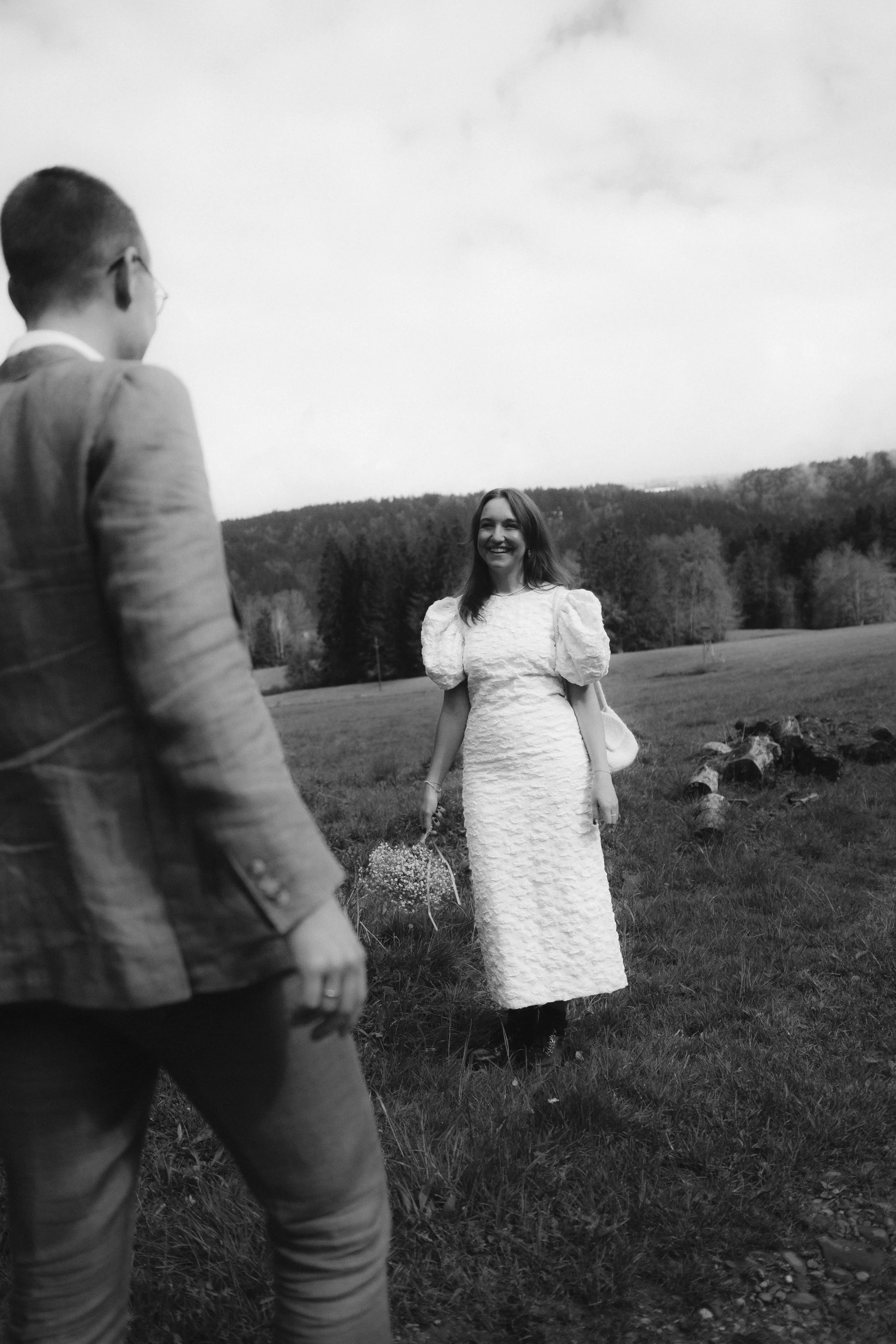 A woman in a long dress and puffed sleeves smiling at a man in a suit on a grassy field with trees and hills in the background.