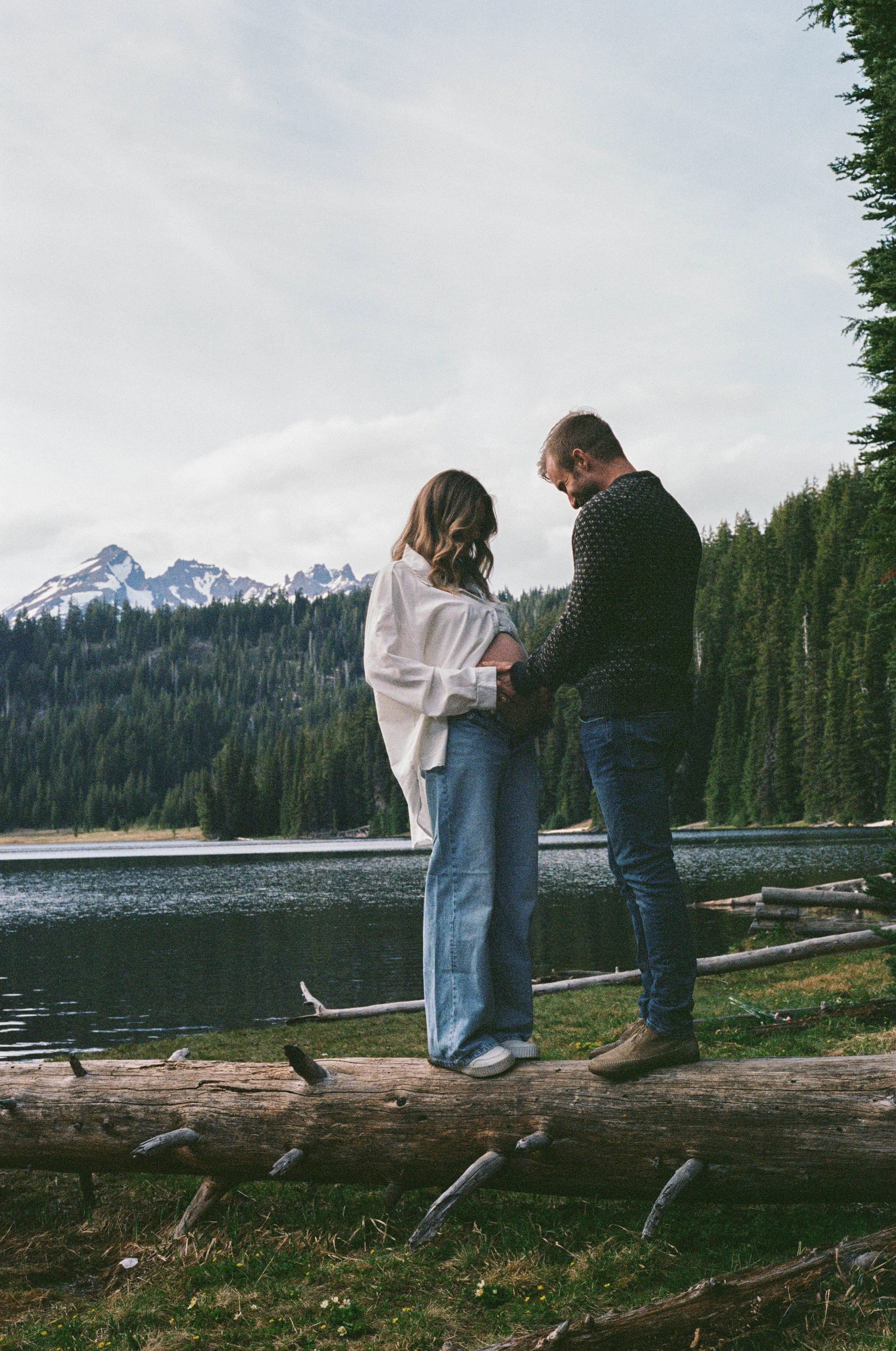 A man and woman standing close together outdoors, holding hands and looking at each other, near a body of water with a backdrop of forested mountains and cloudy sky in Bend Oregon. 