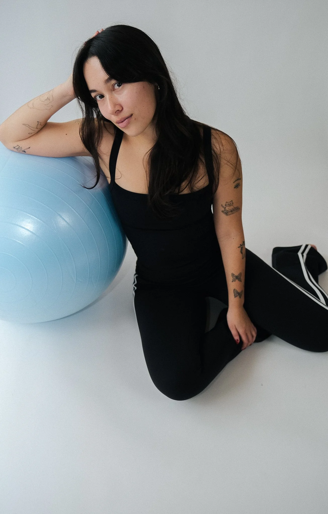 A woman with black hair wearing a black tank top and black leggings, sitting on the floor beside a large blue exercise ball, with one arm resting on the ball and the other on her knee, in a minimalistic room with a plain white background.