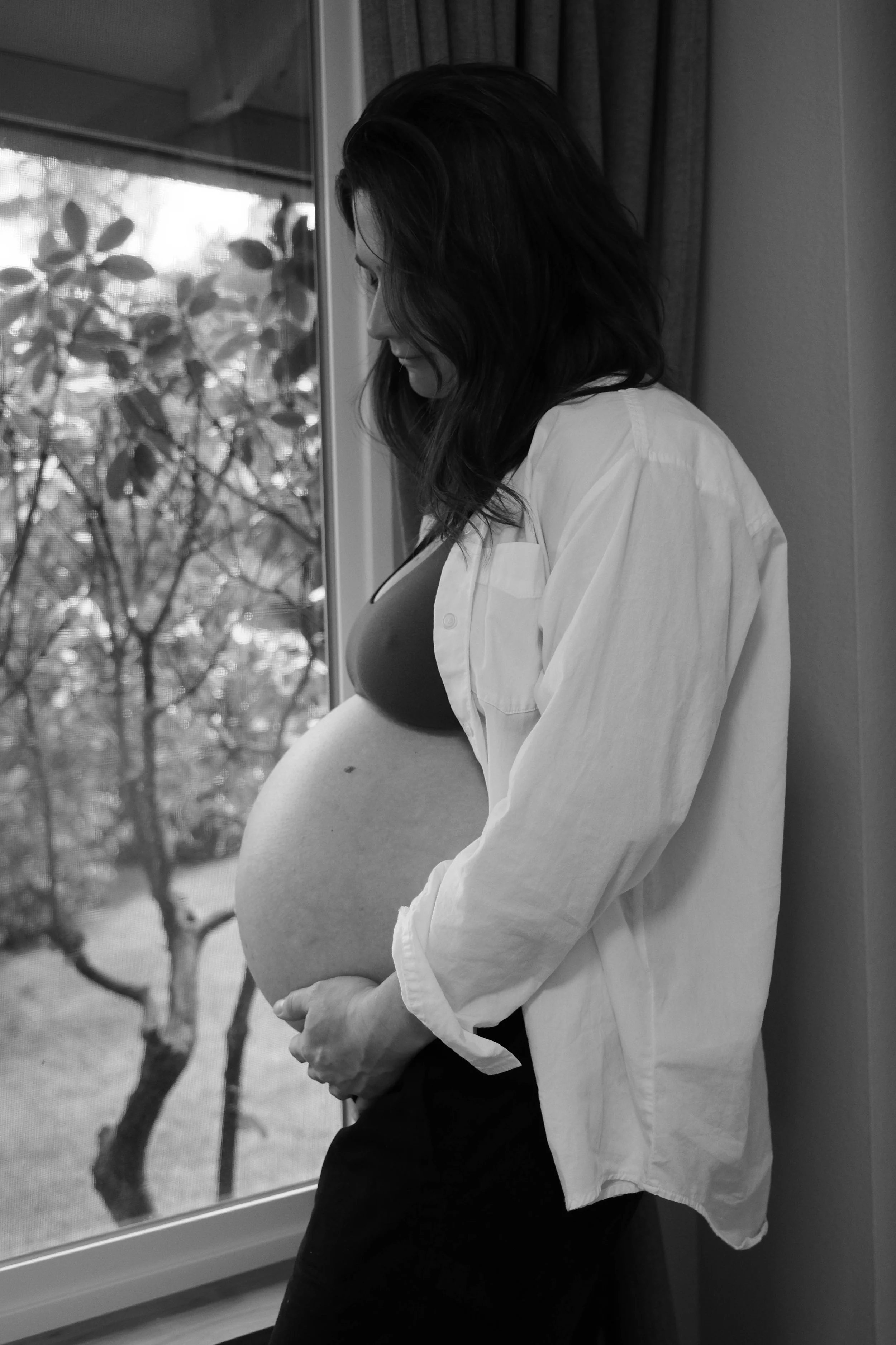 A pregnant woman with long dark hair standing by a window, looking down, wearing a white shirt and black top.