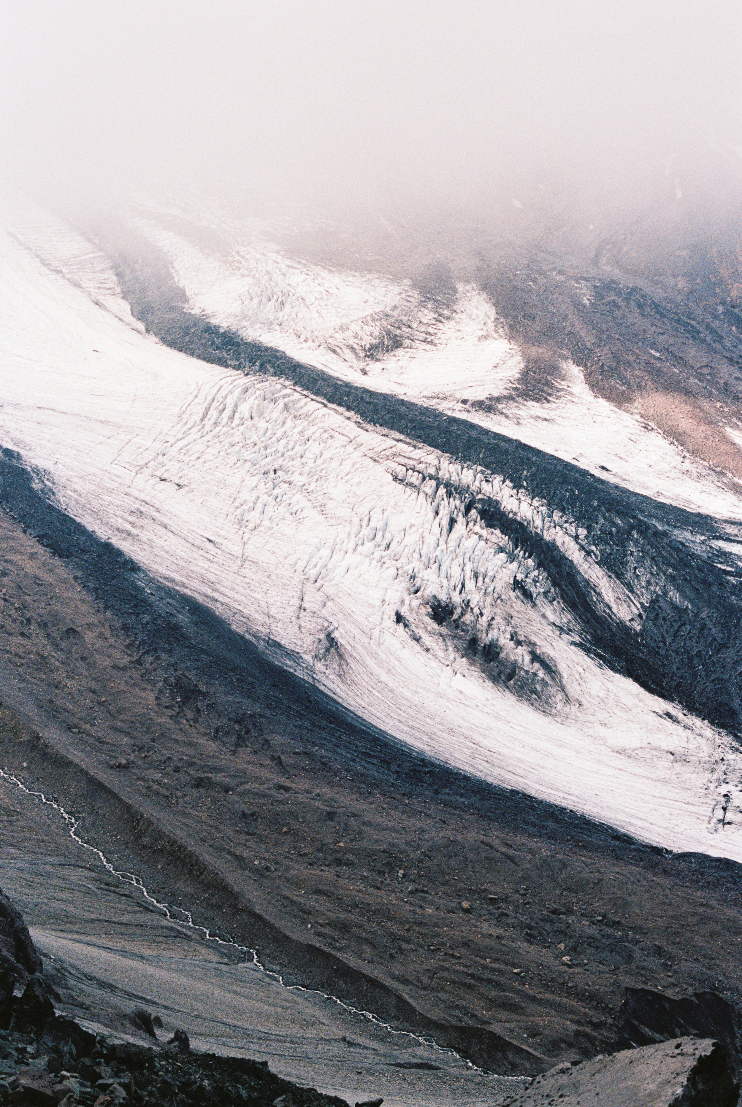 Aerial view of a glacier with melting ice, dark rocky terrain, and a small river flowing at the bottom.