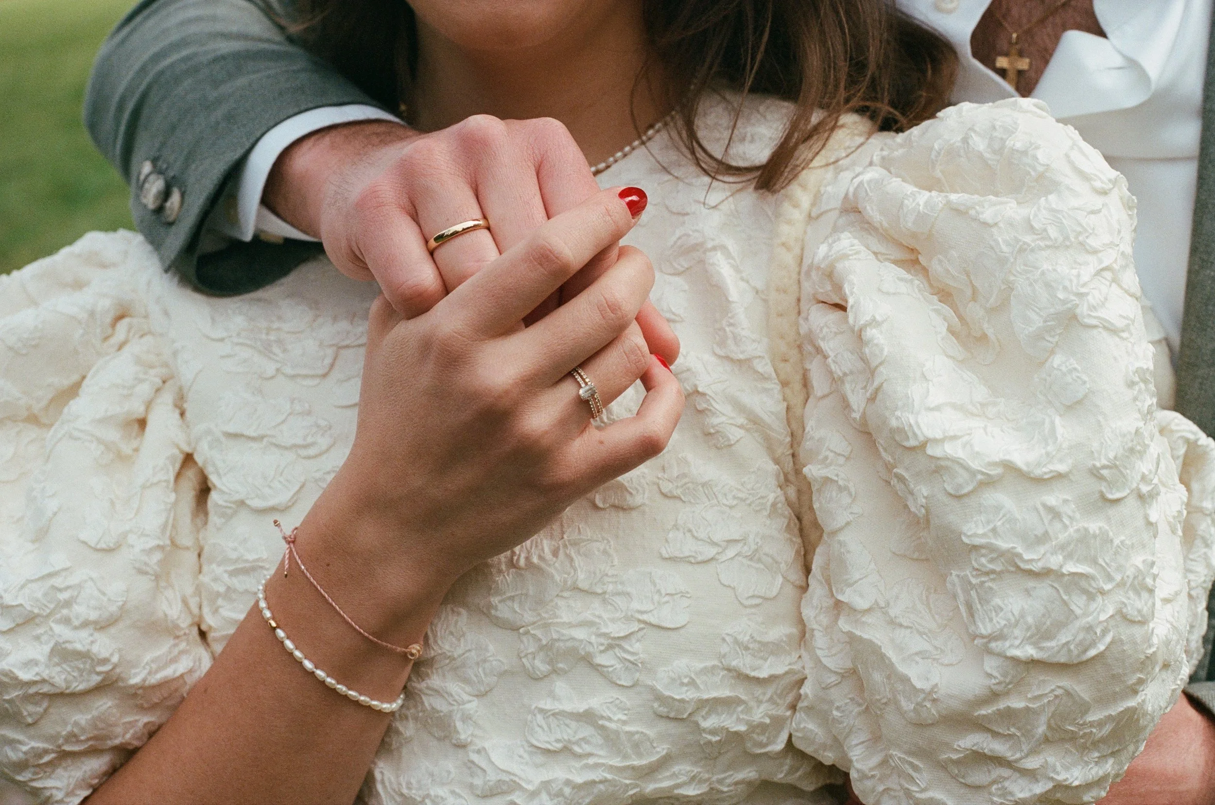 Close-up of a couple's hands intertwined, showing wedding rings. The woman is wearing a pearl bracelet and a gold ring, and the man is wearing a wedding band. The woman is dressed in a textured white wedding dress with lace and puffed sleeves.