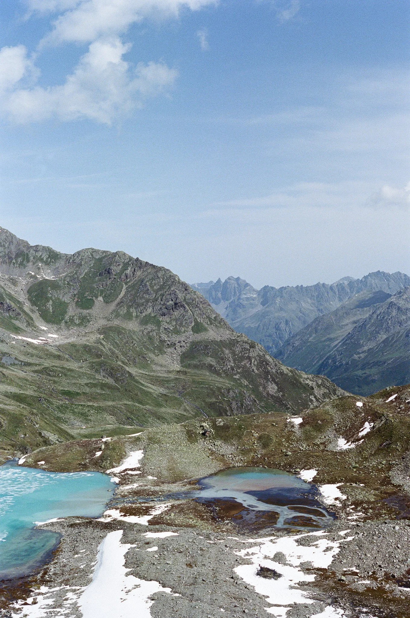Mountain landscape with rocky peaks, patches of snow, and small lakes under a partly cloudy sky.