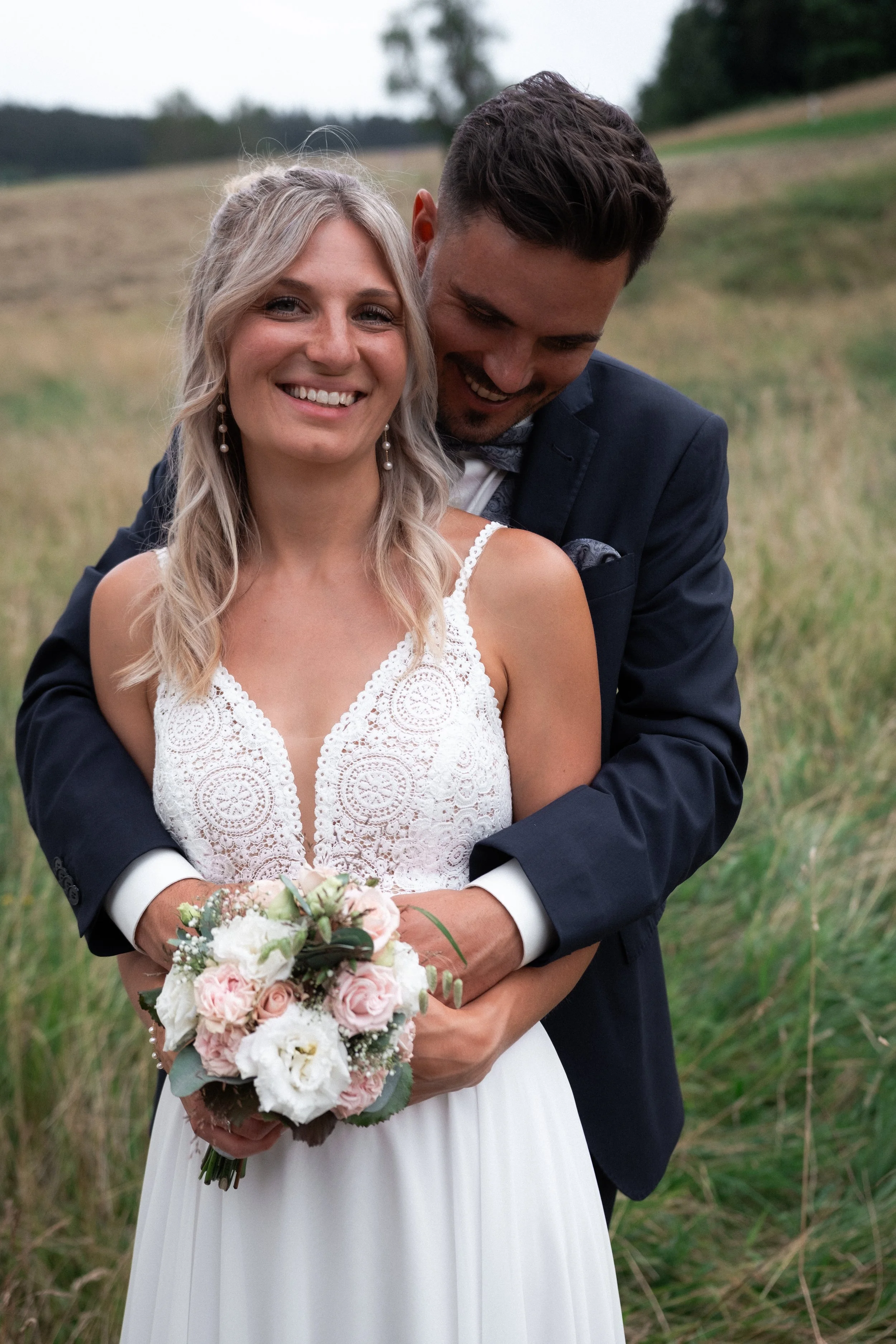 A smiling bride holding a bouquet of pink and white roses, standing in an outdoor field with a groom hugging her from behind.