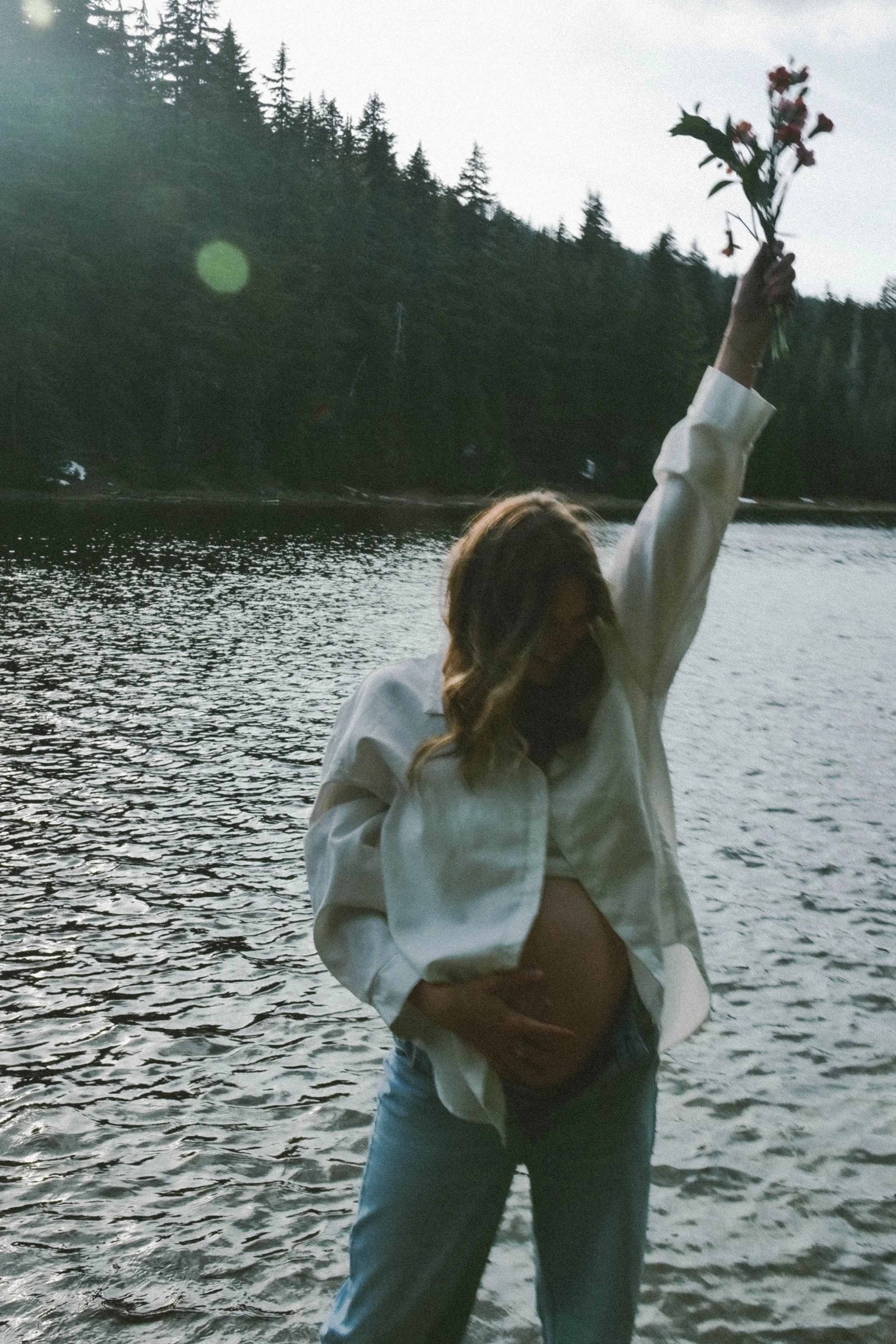 A woman stands near a body of water holding a bunch of flowers in her raised hand during sunset or dusk, with a lush, forested hillside in the background in Oregon, Bend. 