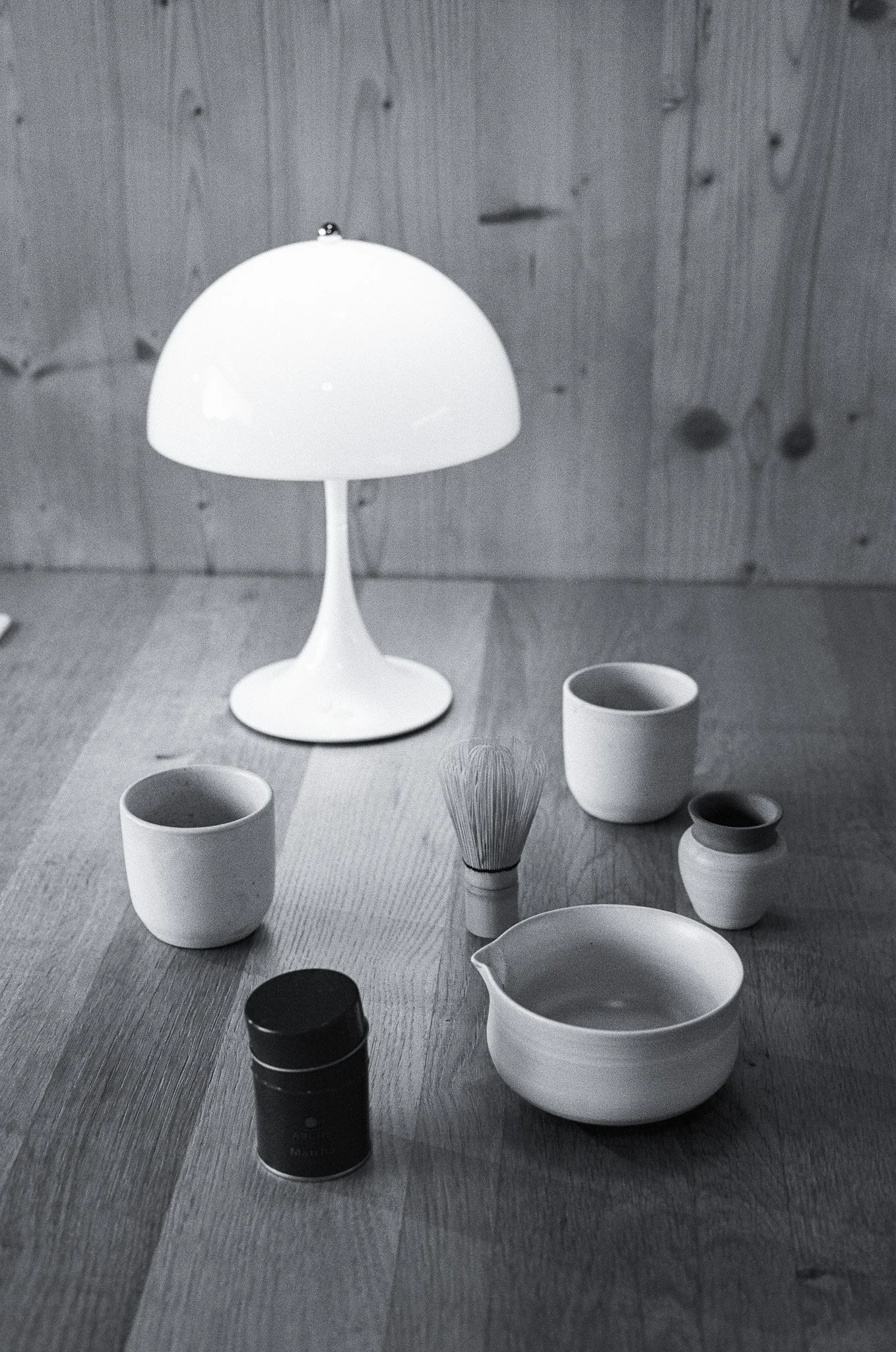 A collection of ceramic cups and bowls, a tea whisk, and a small black container on a wooden table illuminated by a modern white tabletop lamp against a wooden wall background.