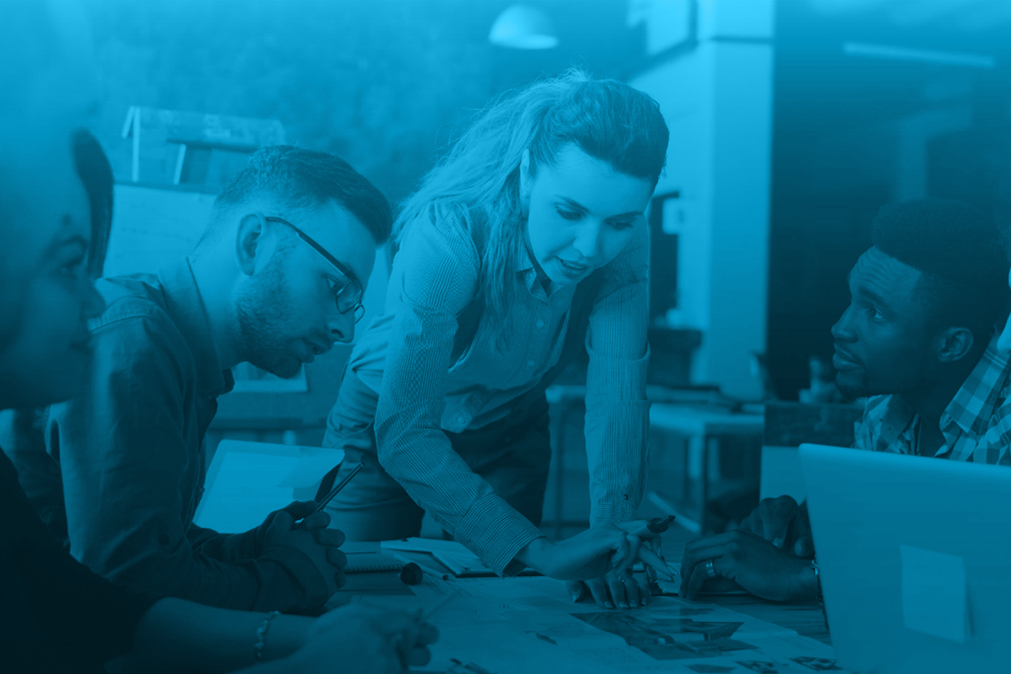 Group of five diverse people in a business meeting, engaged in discussion, with papers and a laptop on the table, in an office setting.