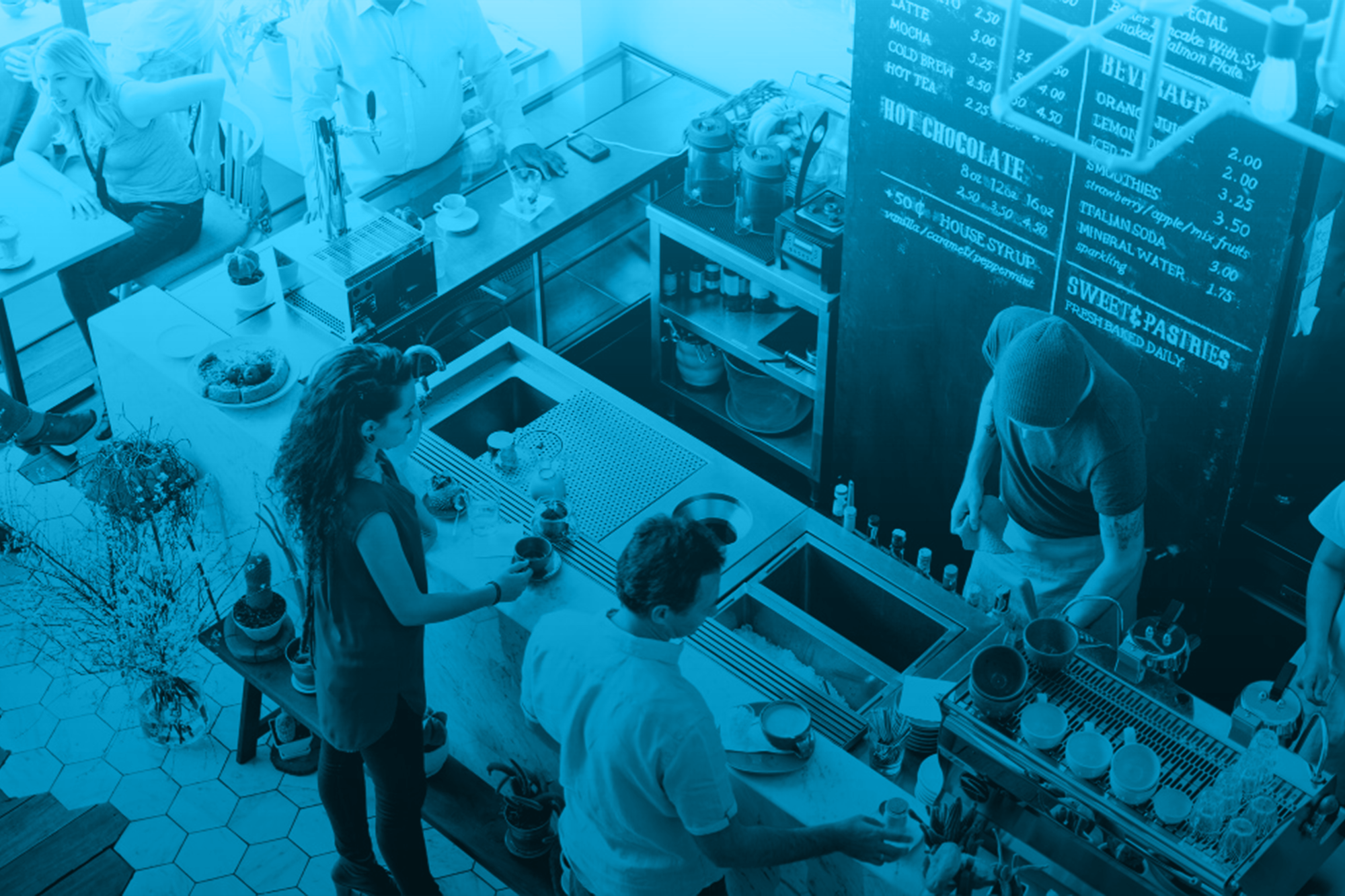 Interior of a cafe showing customers ordering and baristas preparing drinks at a counter, with a chalkboard menu listing various beverages and prices.