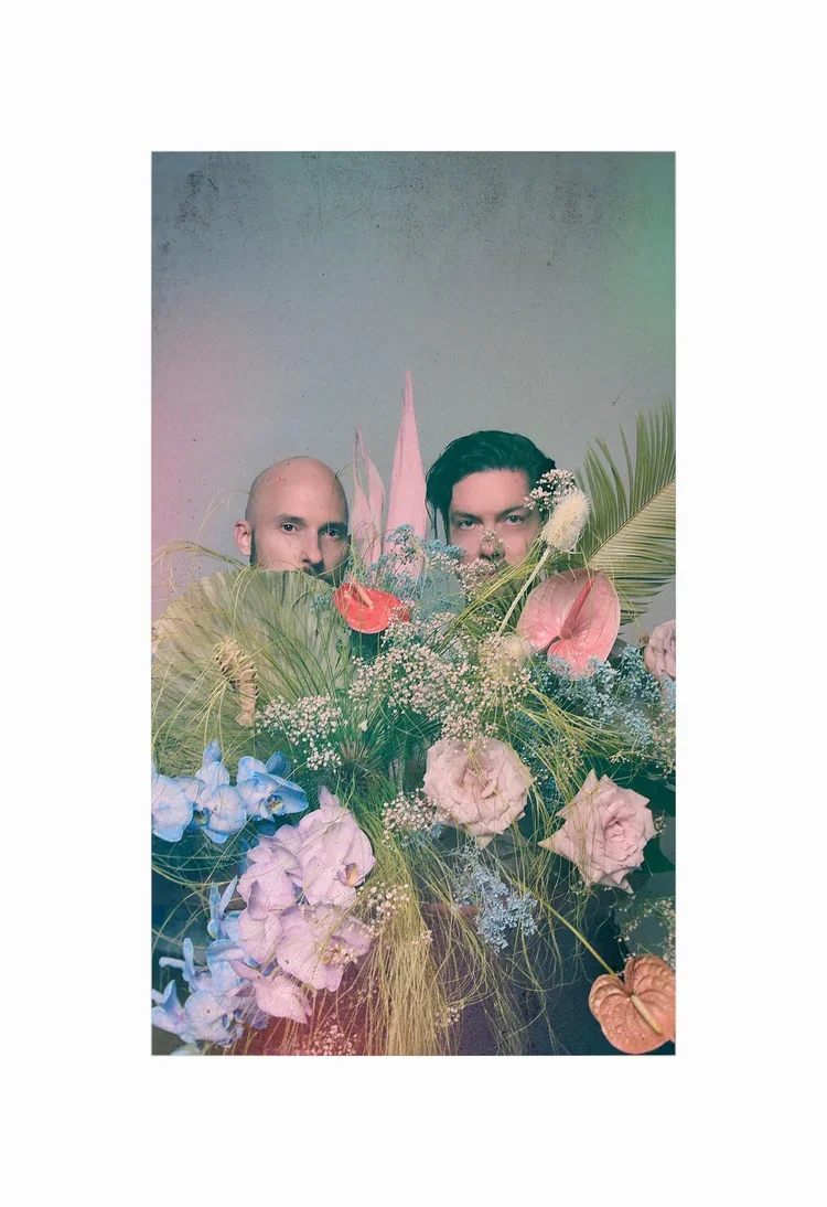 Two men holding a large bouquet of pink and purple flowers with green leaves.