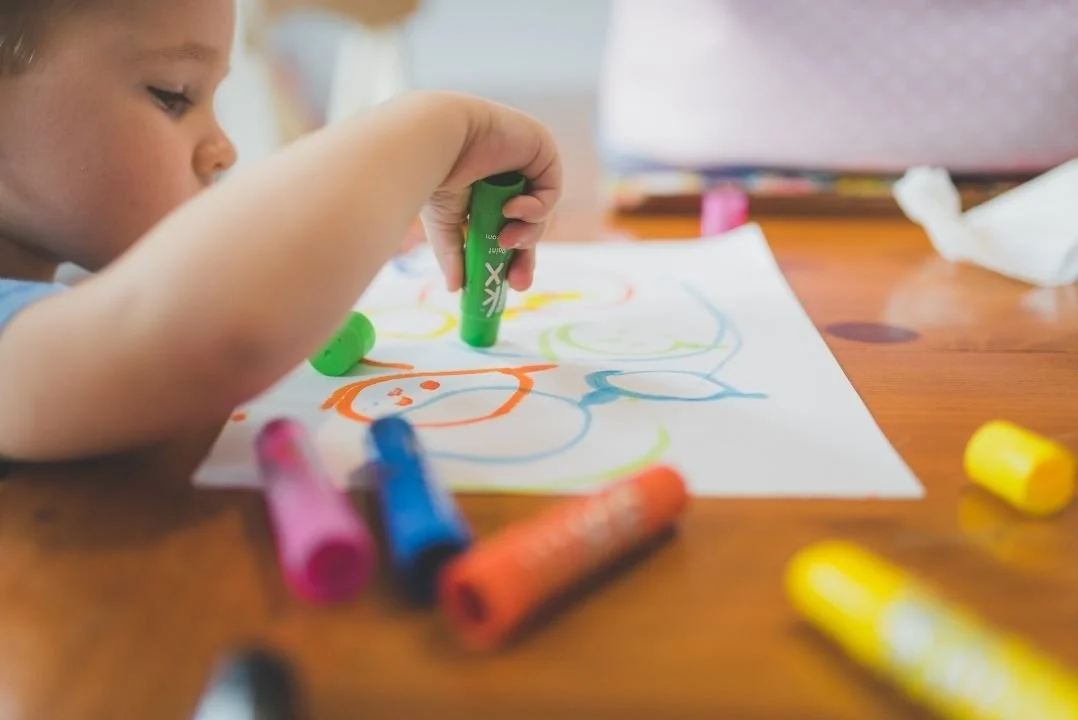 Child drawing colorful shapes with markers on paper, surrounded by markers on a wooden table.