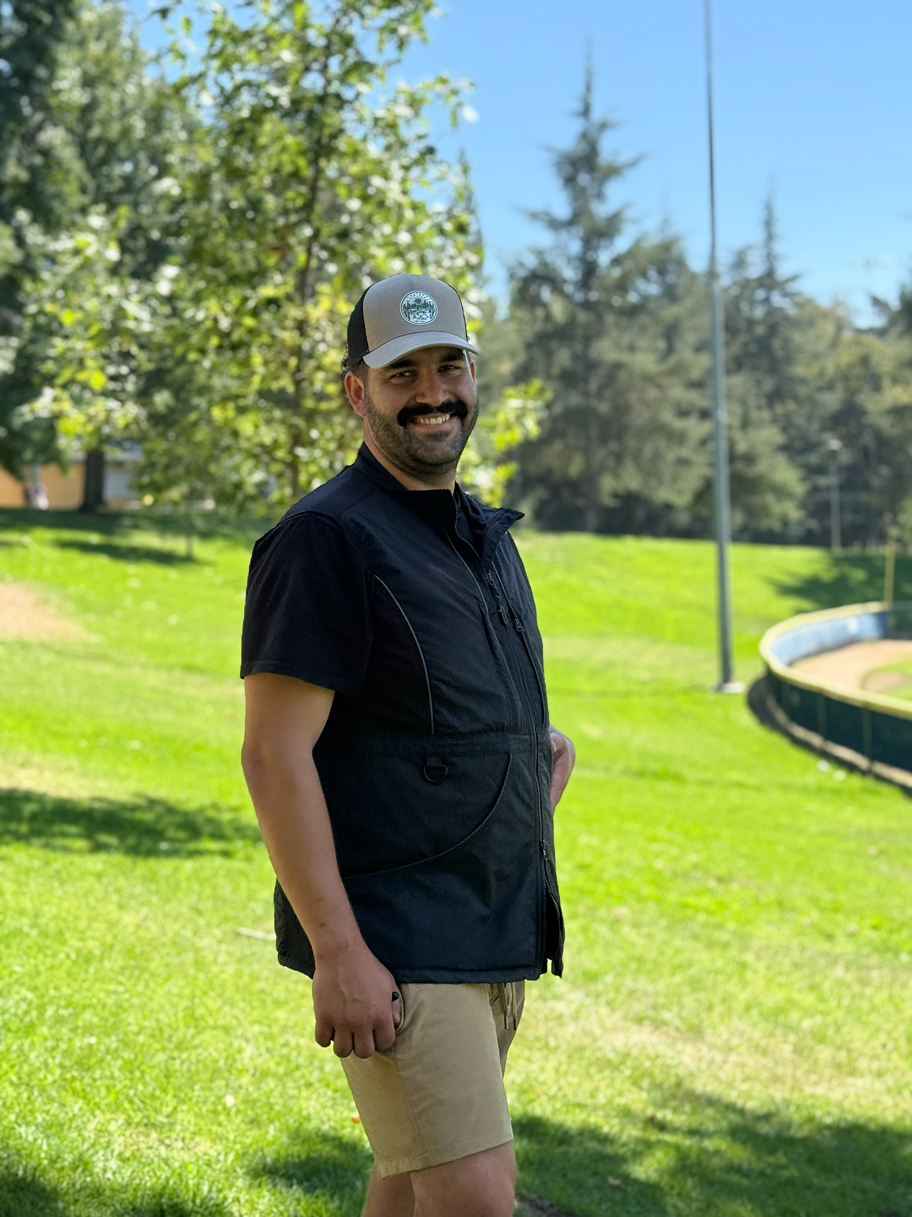 A man with a beard and mustache smiling outdoors, wearing a gray and black cap and a black jacket, standing on a grassy area with trees in the background on a sunny day.