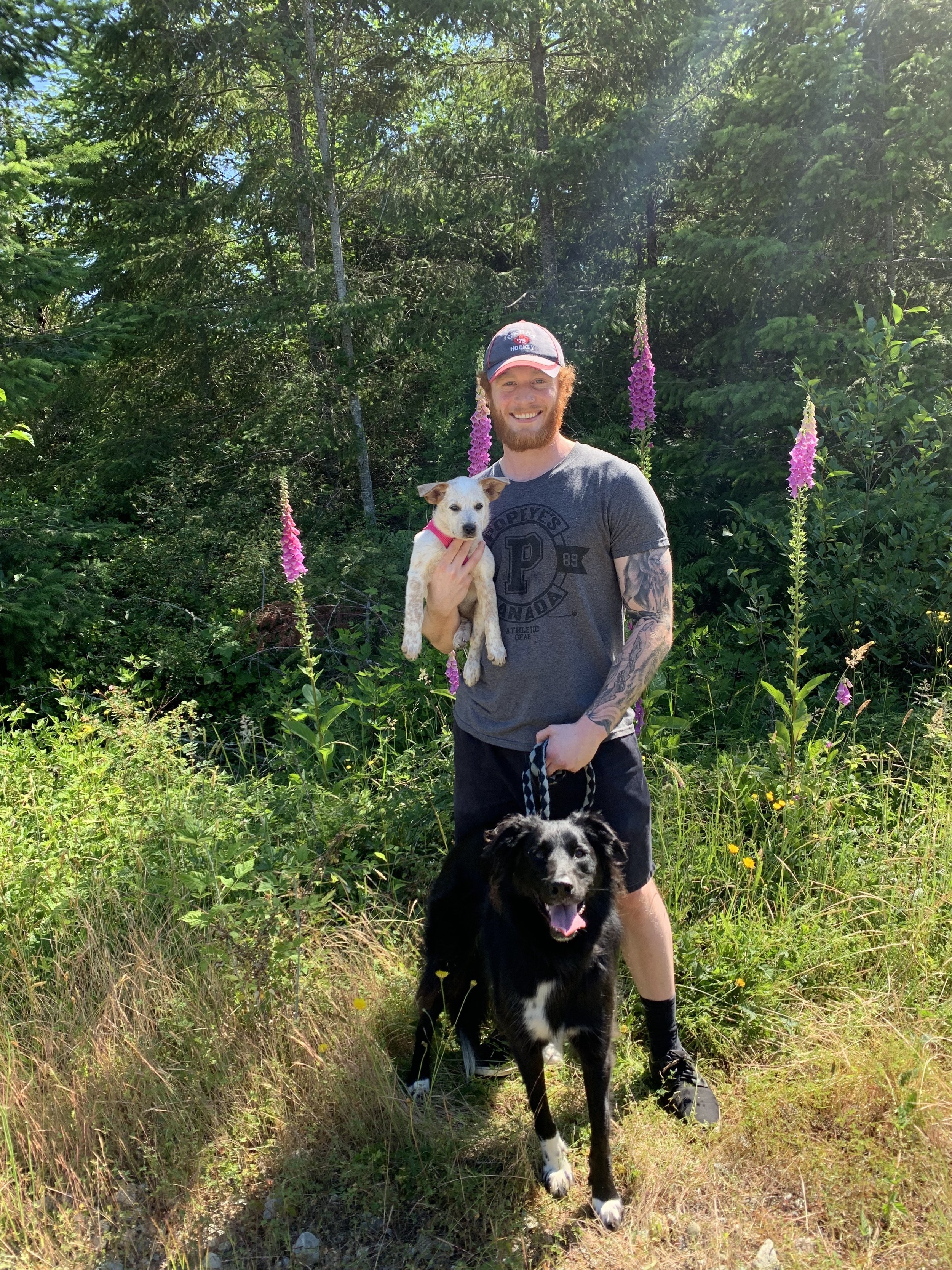 A man with a beard and tattooed arm smiling and holding a small white dog, standing outdoors among green trees and pink flowers, with a black dog on a leash in front.