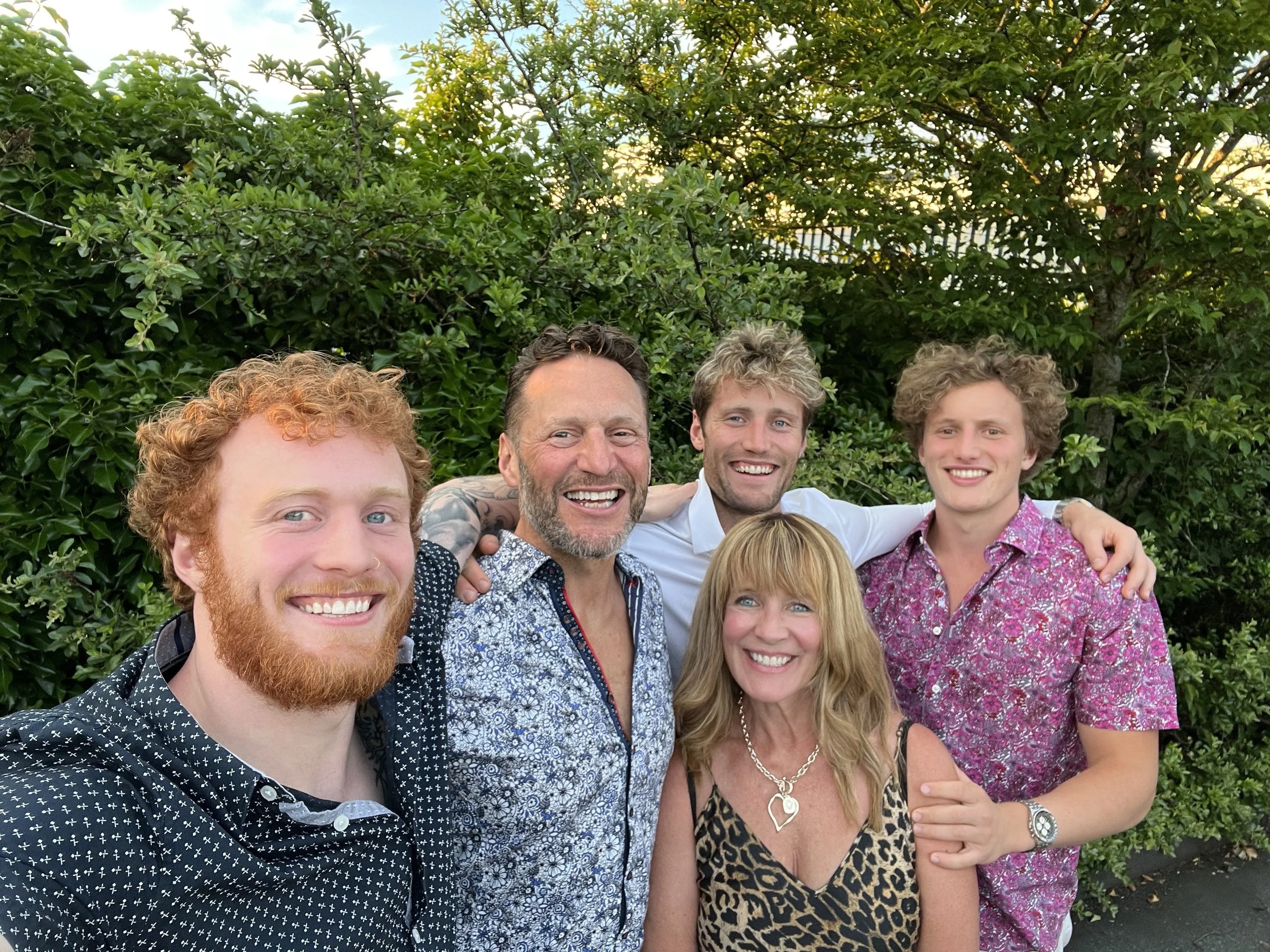 six people smiling and posing for a photo outdoors in front of green bushes, with three men and one woman in the front row and one man and one woman in the back row.