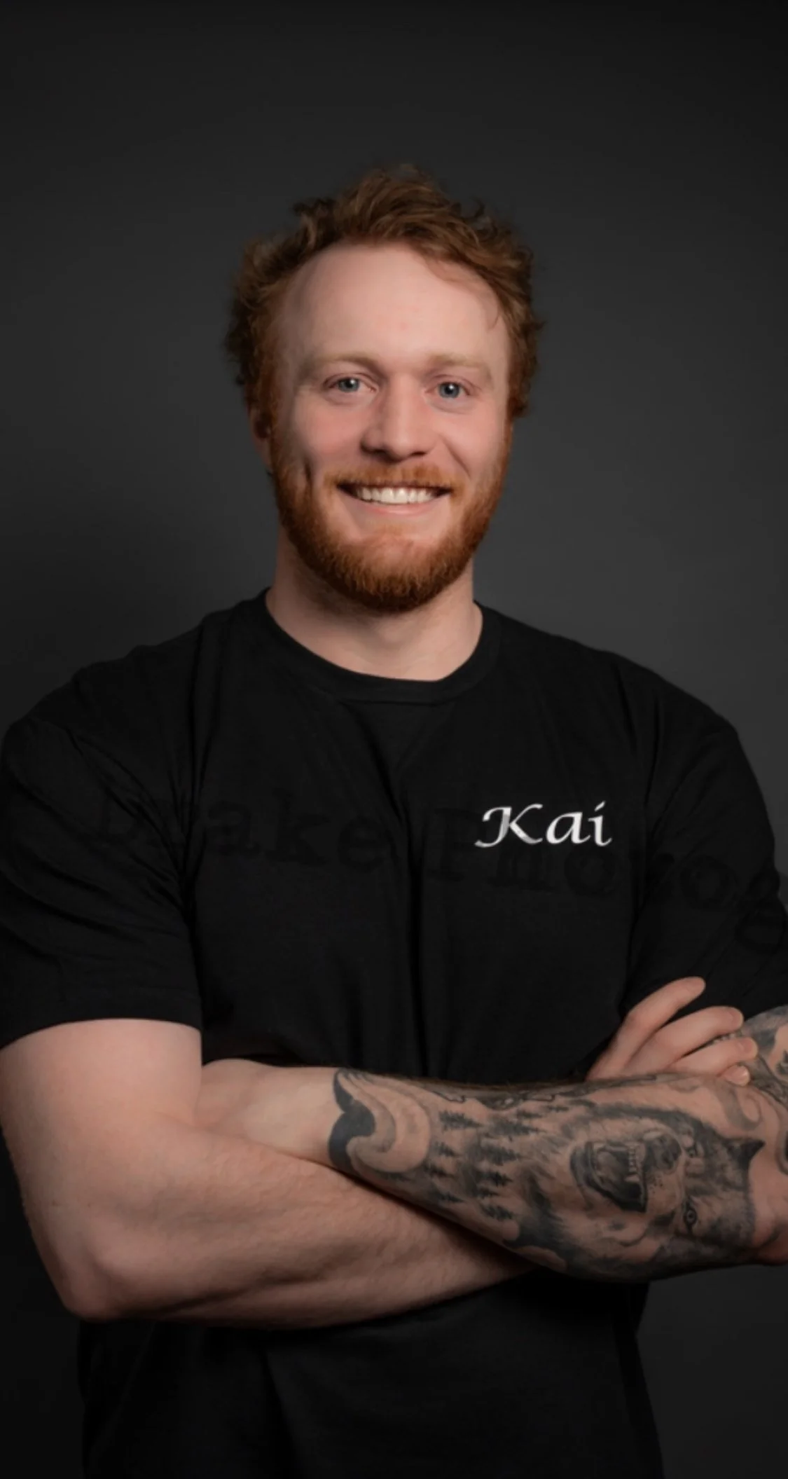 A smiling man with red hair and a beard, wearing a black t-shirt with the name 'Kai' on it, standing against a dark background.