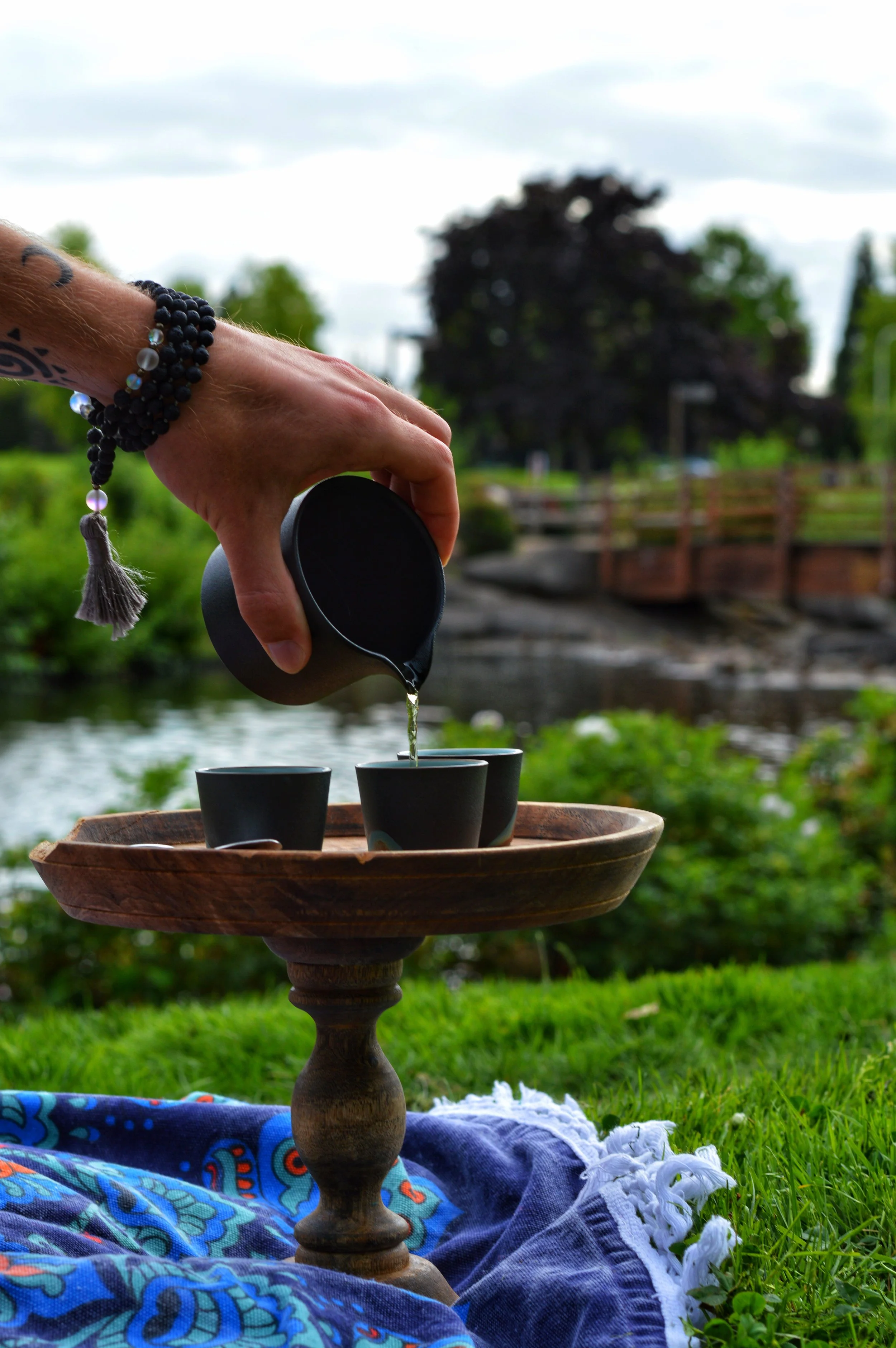 A person pouring tea from a small black teapot into black cups on a wooden stand outdoors near a river, with green grass, shrubs, and trees in the background.