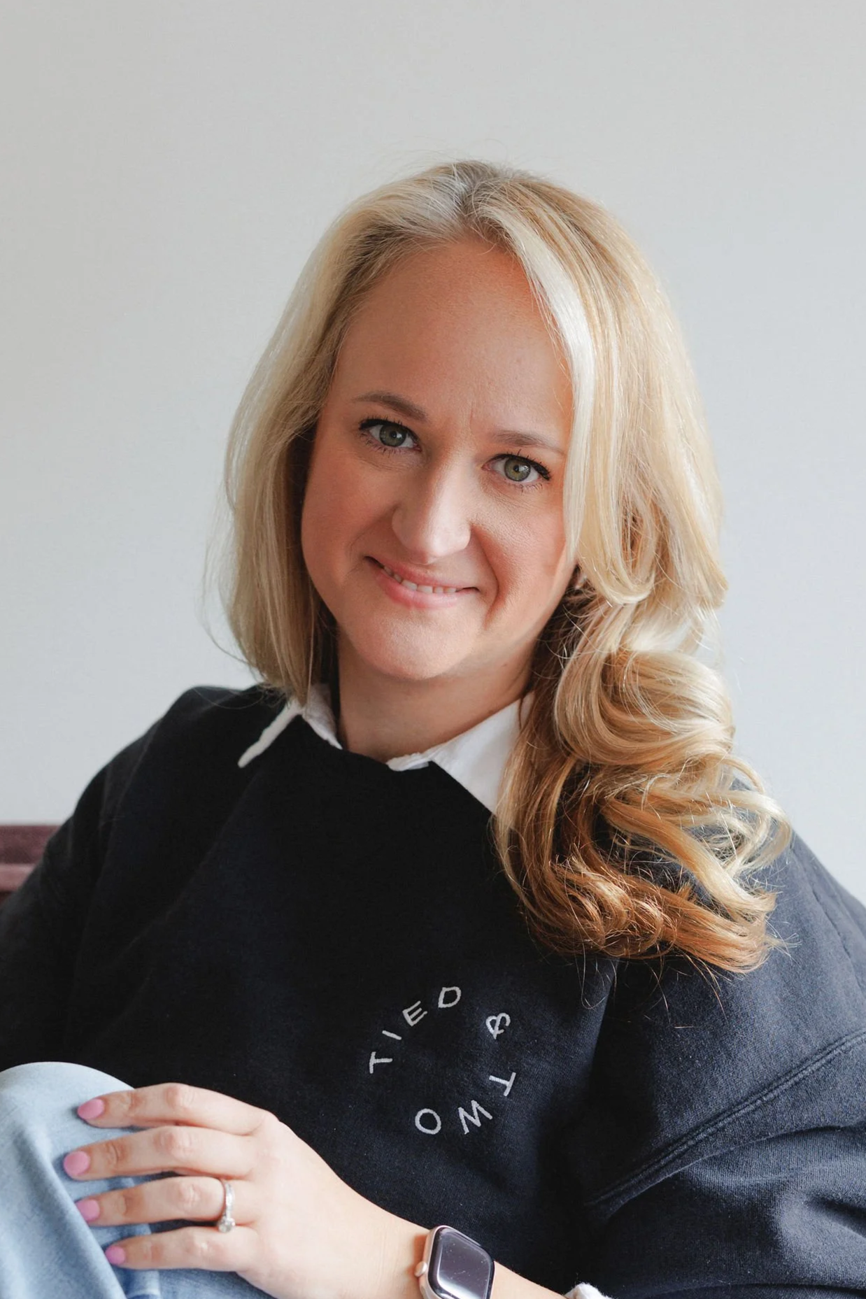 A woman with long blonde wavy hair, smiling, wearing a black shirt against a plain light gray background.