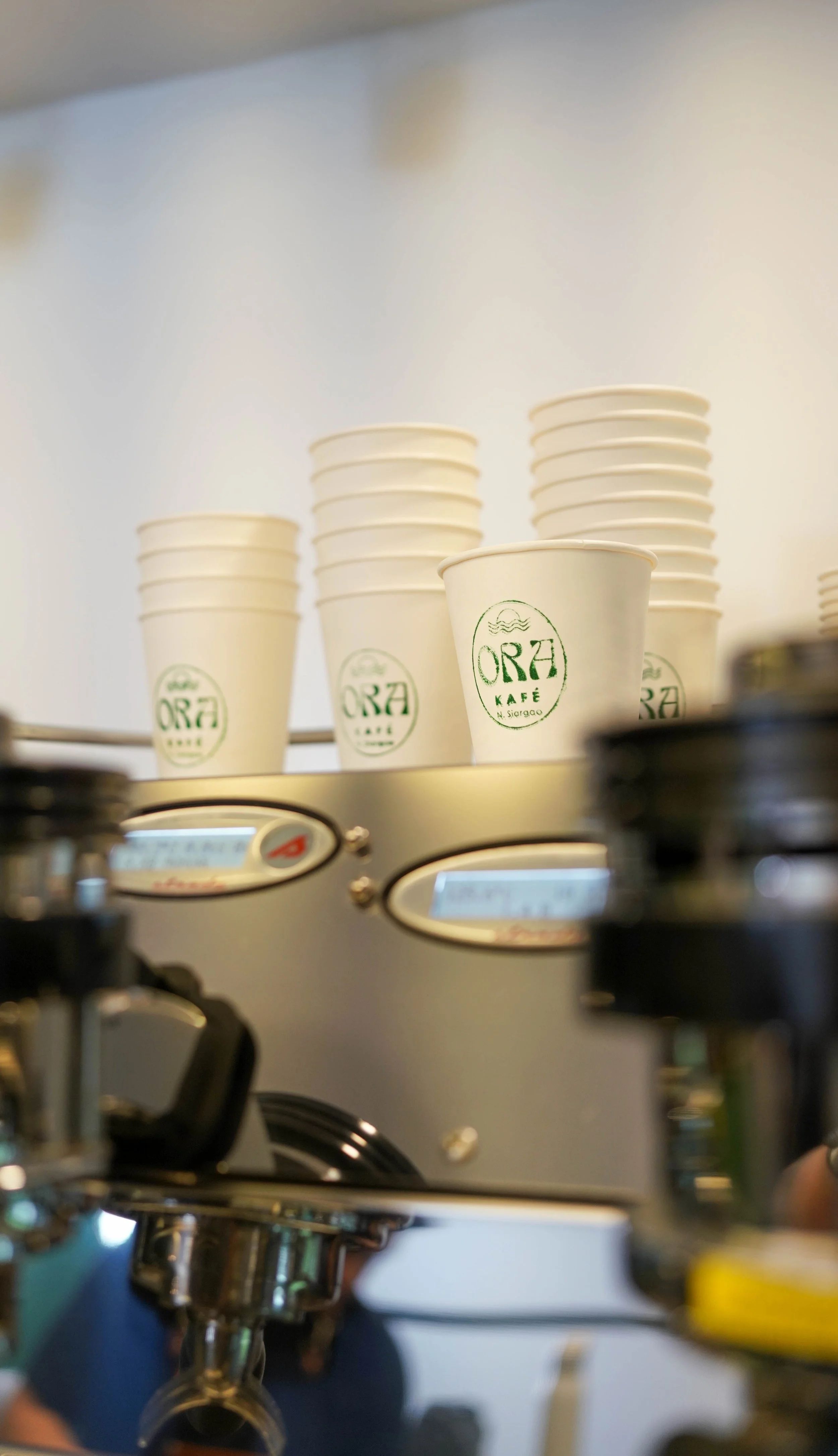 Stack of white paper coffee cups on a coffee machine with the logo 'ORA KAFE'.