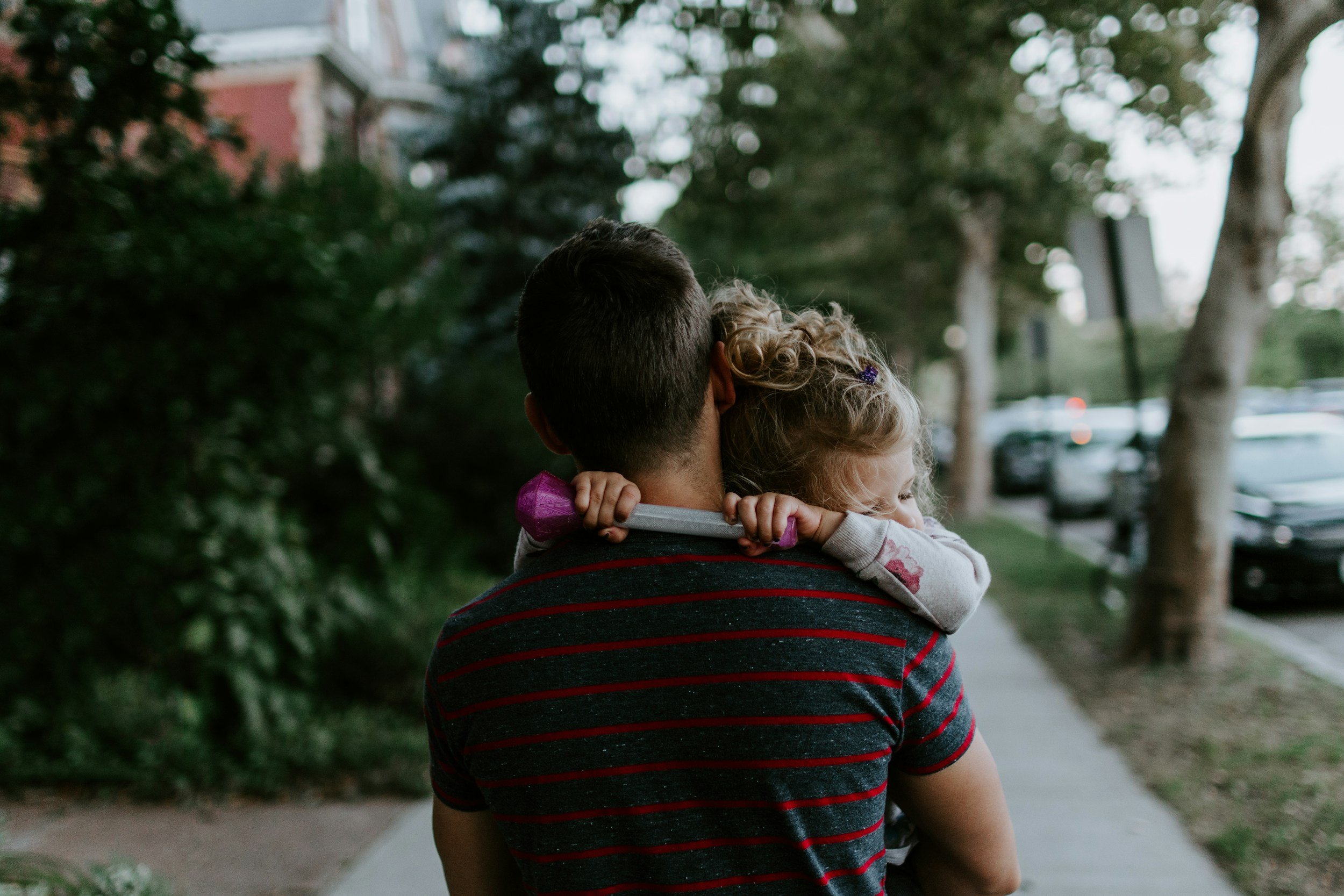 A person carrying a young girl on their shoulders on a sidewalk lined with trees and parked cars.
